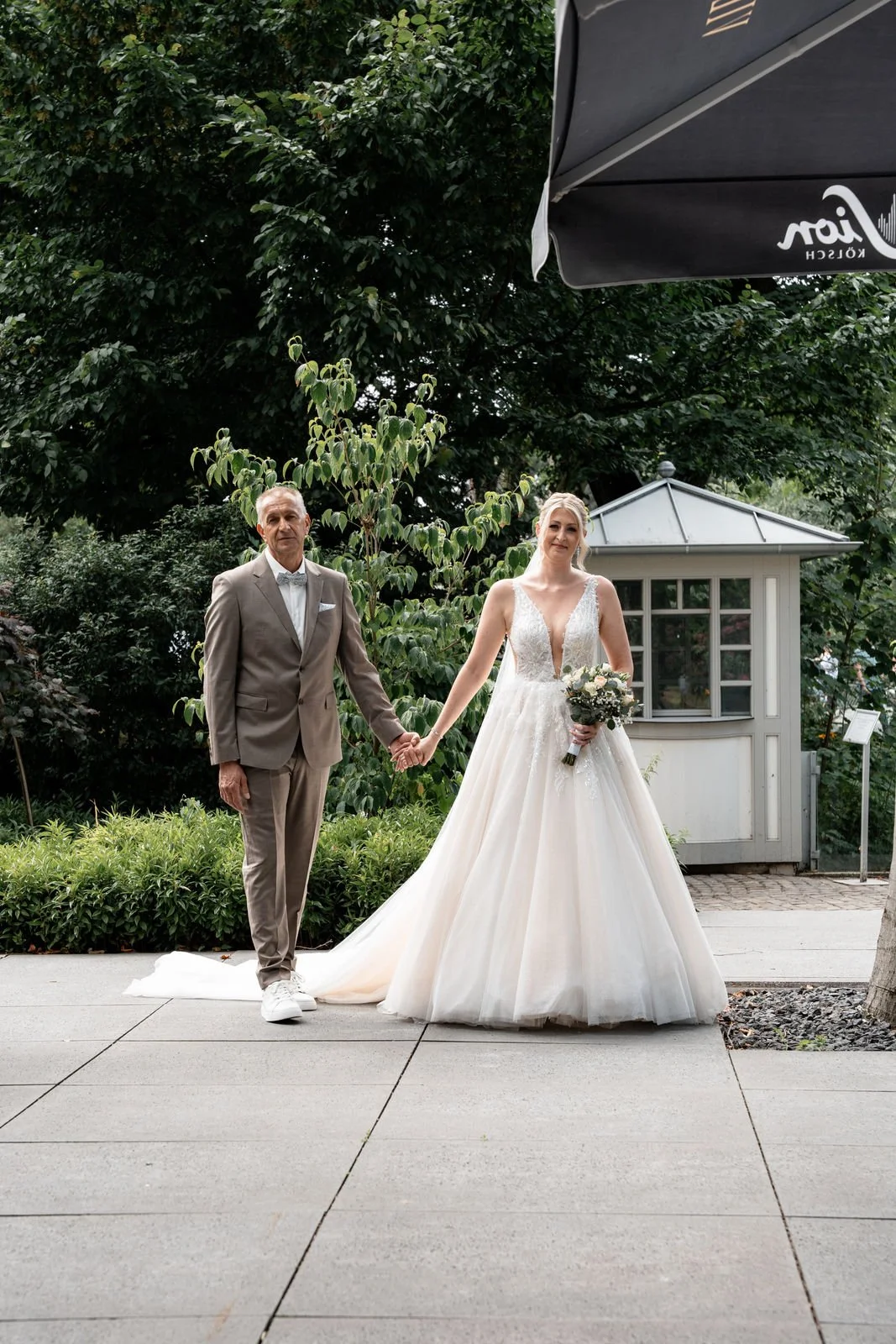 Braut und älterer Mann, vermutlich Vater, stehen im Freien auf Gehweg, Hand in Hand. Braut trägt weißes Traumhochzeitskleid und hält einen Blumenstrauß. Im Hintergrund sind Bäume und eine kleine weiße Gartenlaube sichtbar.