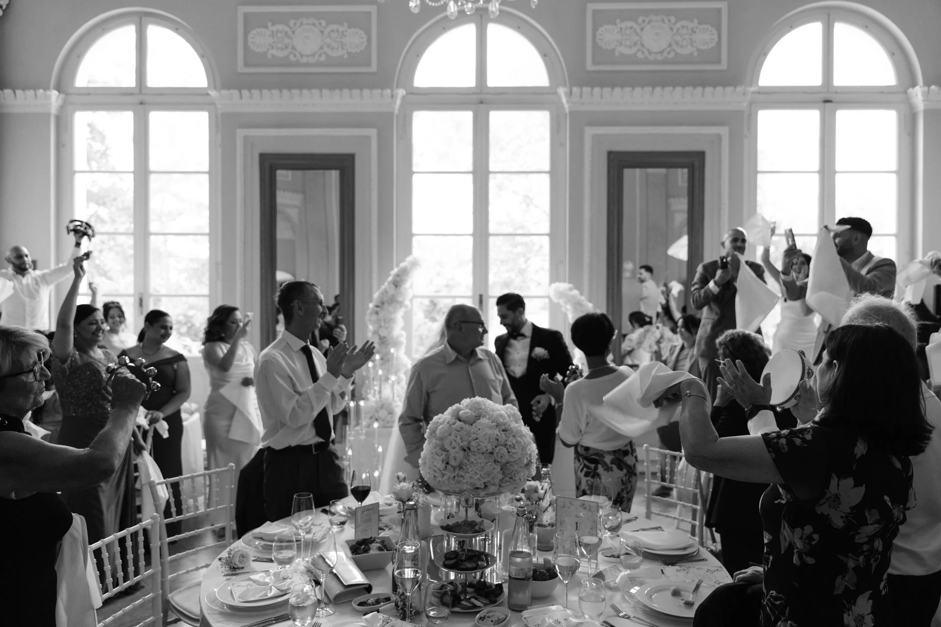Ein feierlicher Hochzeitsempfang in einem eleganten Saal mit vielen Gästen, die feiern, applaudieren und Fotos machen. Dekorative Blumen und festliche Tafel sind sichtbar.