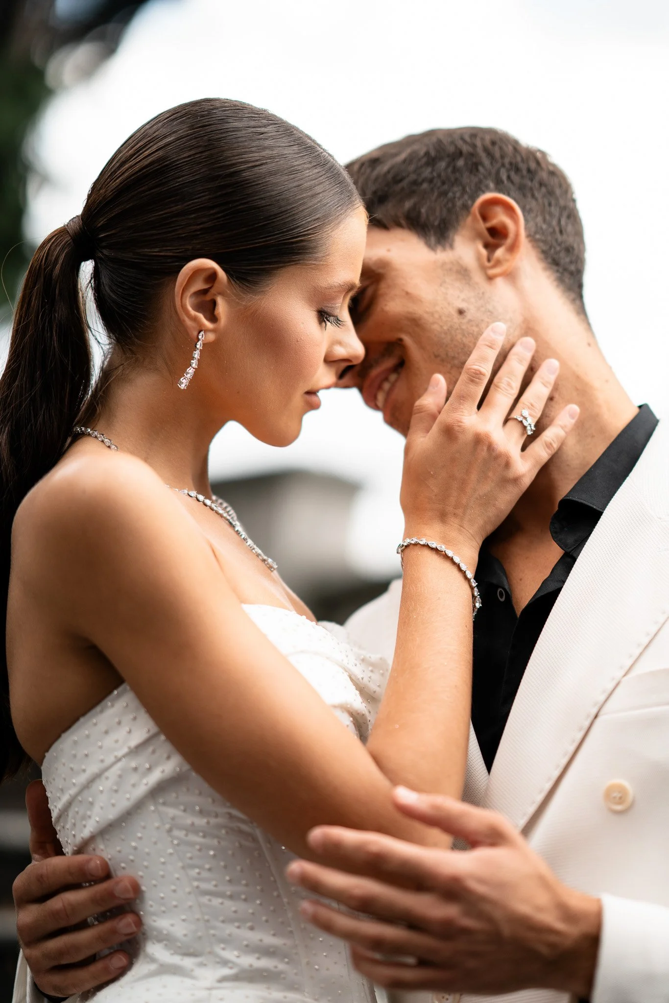 Ein Brautpaar in emotionalem Moment bei ihrer Hochzeit, eng umarmt und sich an die Stirn blickend, mit eleganter Kleidung und Schmuck, outdoors an einem grauen Tag.