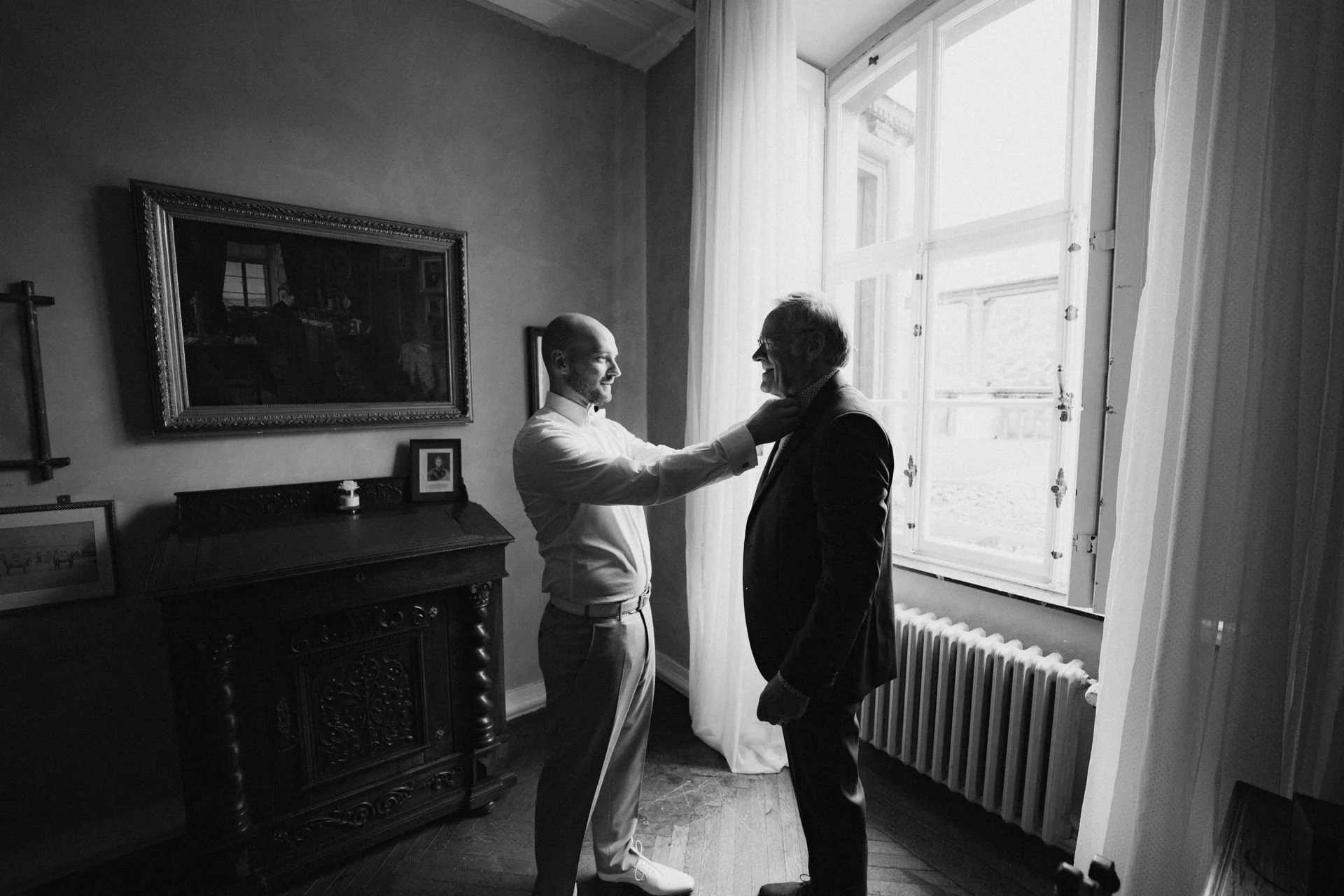 Zwei Männer, vermutlich ein Vater und sein Sohn, stehen im Raum bei einem herzlichen Moment, beide lächelnd. Der Jüngere trägt helle Kleidung, der Ältere einen Anzug.