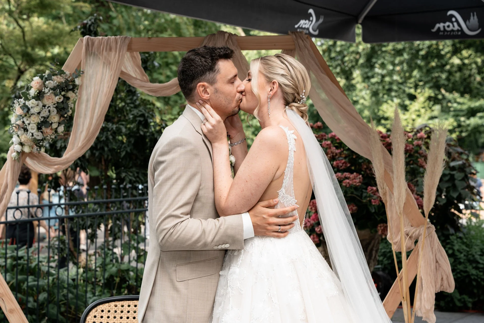 Ein Brautpaar teilt einen Kuss während ihrer Hochzeit im Freien, vor einem dekorativen Holzbogen mit Blumen und Tüchern, umgeben von Bäumen.