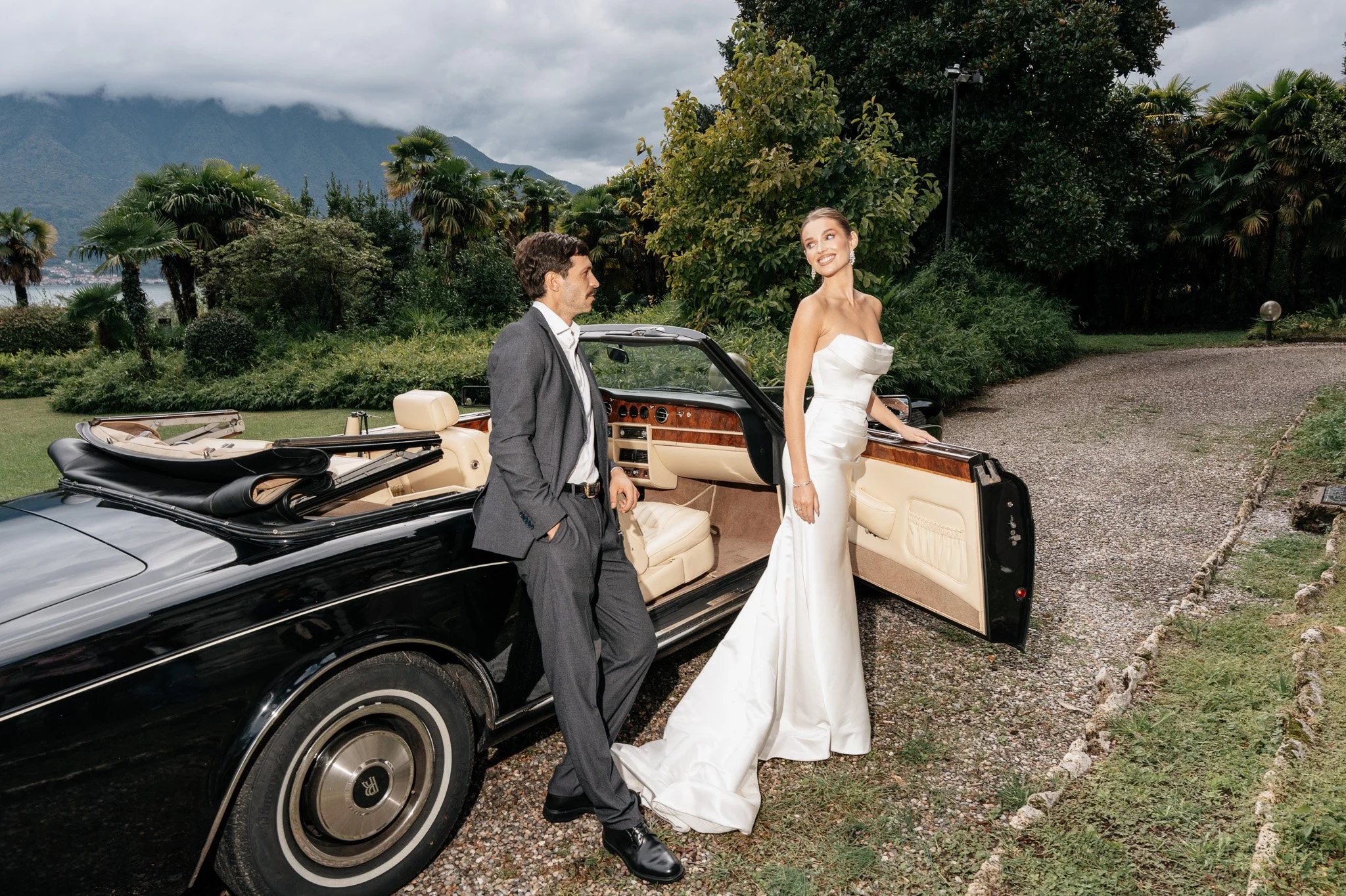 Ein Mann in Anzug und eine Frau in einem weißen Hochzeitskleid stehen neben einem schwarzen Cabriolet auf einer grüne Wiese vor einer Anlage mit Bäumen und Bergen im Hintergrund.