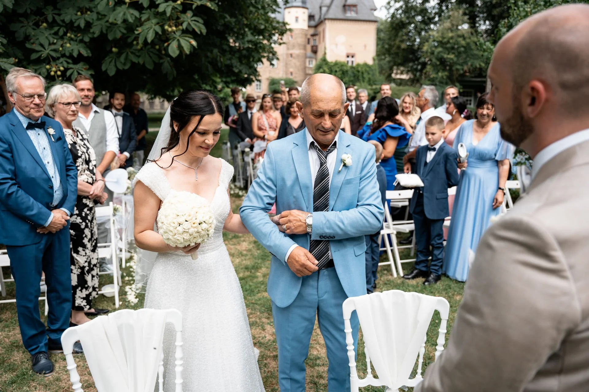 Eine Hochzeit im Freien mit mehreren Gästen, die nebeneinander im Hintergrund stehen. Im Vordergrund sind ein Bräutigam im beige Anzug mit Bart, die Braut in einem weißen Hochzeitskleid, eine ältere Dame und ein älterer Herr im blauen Anzug zu sehen,
