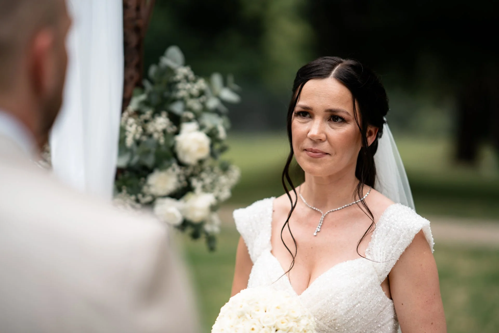Braut mit dunklem Haar trägt ein weißes Hochzeitskleid und sieht den Bräutigam an, im Hintergrund Blumen und grüne Bäume.