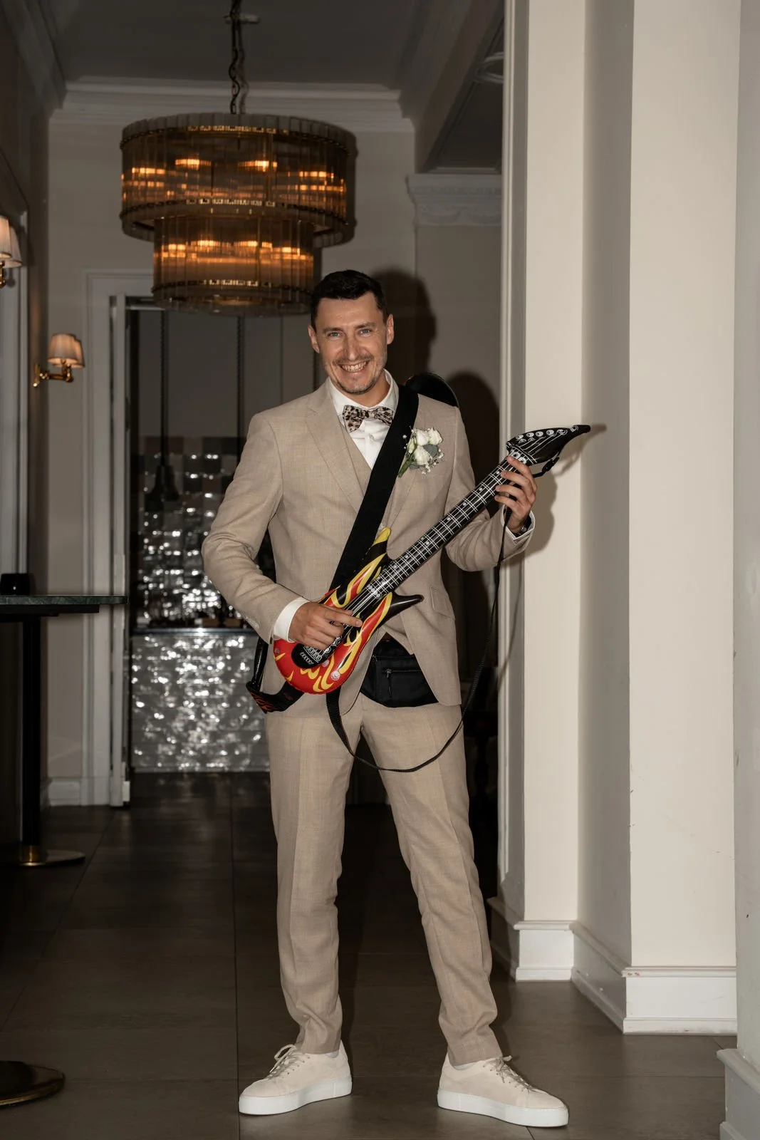 Mann in beige Anzug mit Blumenansteckerei, weißem Hemd, Schleife, weiße Sneaker, hält eine elektrische Gitarre mit Flammenmuster, steht in einem eleganten Raum mit Kronleuchter.