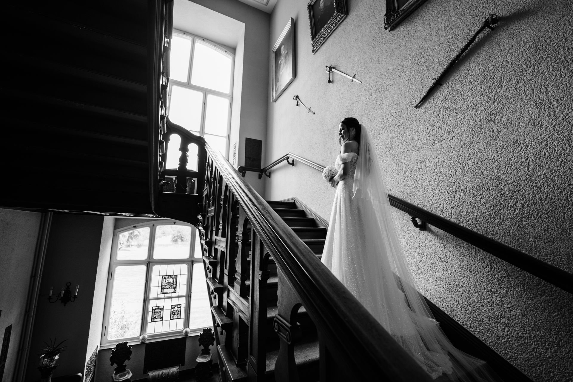 Braut in Hochzeitskleid mit Schleier steht auf einer Treppe im historischen Gebäude, im Hintergrund großes Fenster mit buntem Glas. Schwarz-Weiß-Fotografie.