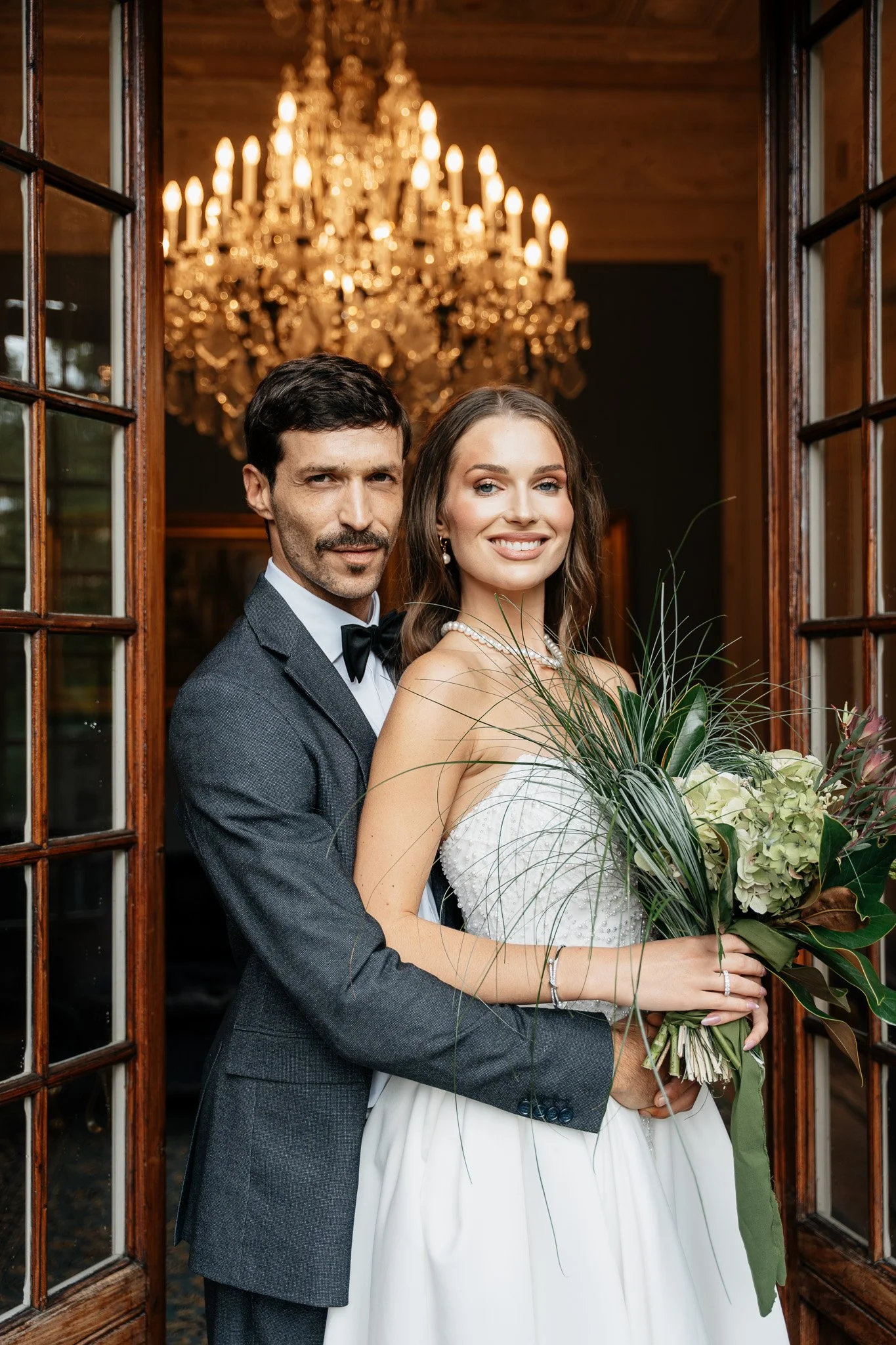 Ein glückliches Brautpaar auf ihrer Hochzeit, standesgemäß mit Blick in die Kamera, vor einer prächtigen Kristalllampe im Hintergrund.