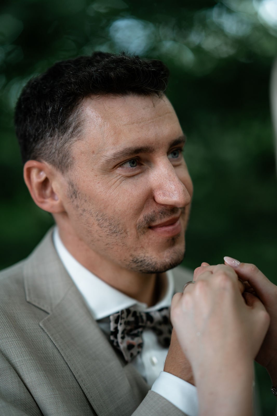 Nahaufnahme eines lachenden Mannes in Anzug mit Fliege, im Freien, mit grüner, unscharfer Baum-Umgebung im Hintergrund.