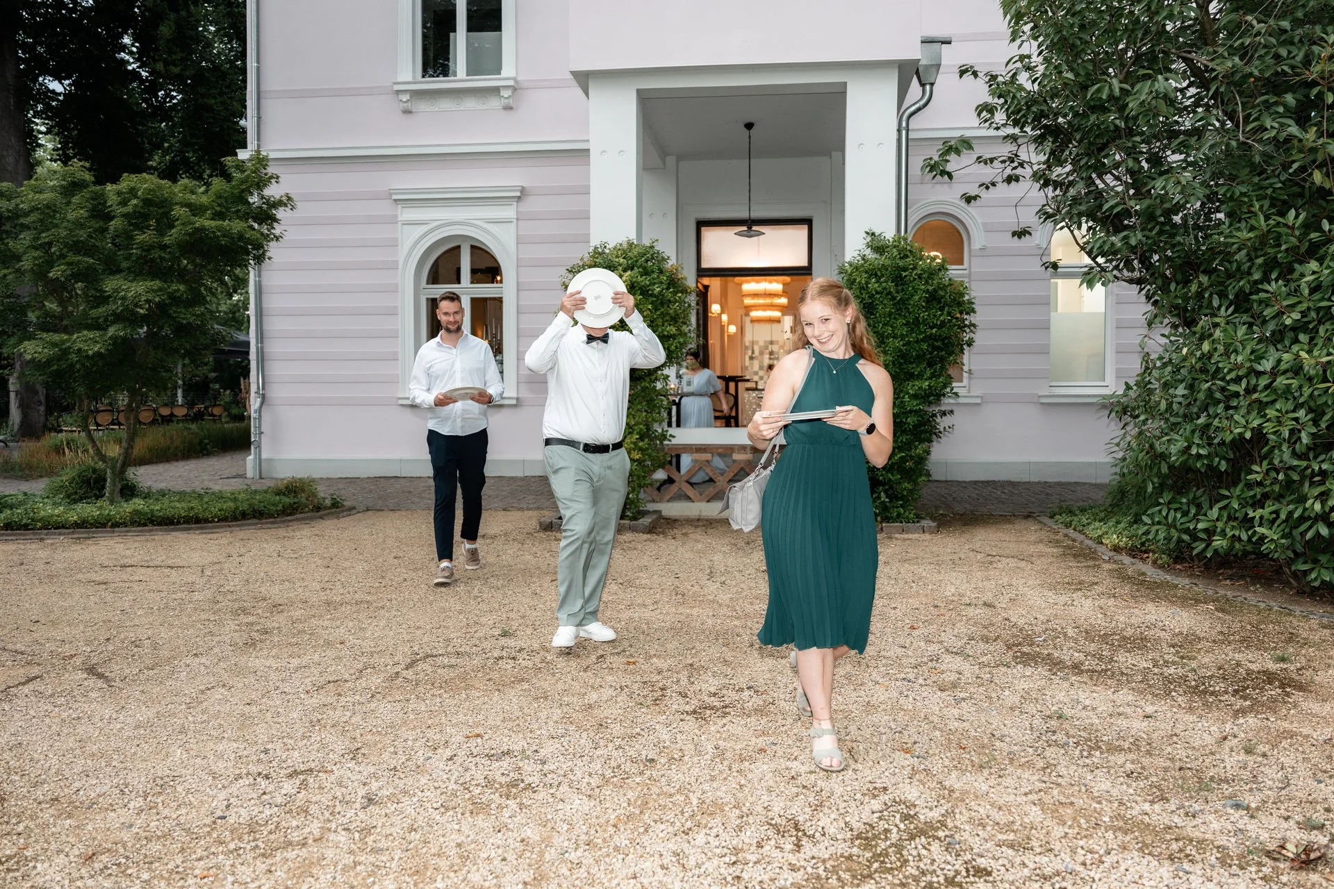 Menschen verlassen ein elegantes Gebäude, darunter eine Frau in einem grünen Kleid, die ein Tablet hält und lächelnd geht, während zwei Männer hinter ihr eine Speisekarte und Teller tragen.