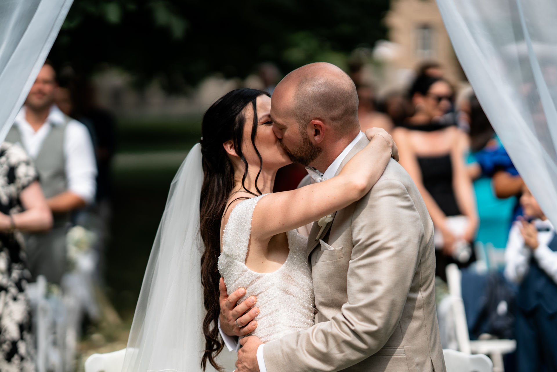 Braut und Bräutigam küssen sich bei ihrer Hochzeit unter einer Gartenlaube, im Hintergrund feiern Gäste.
