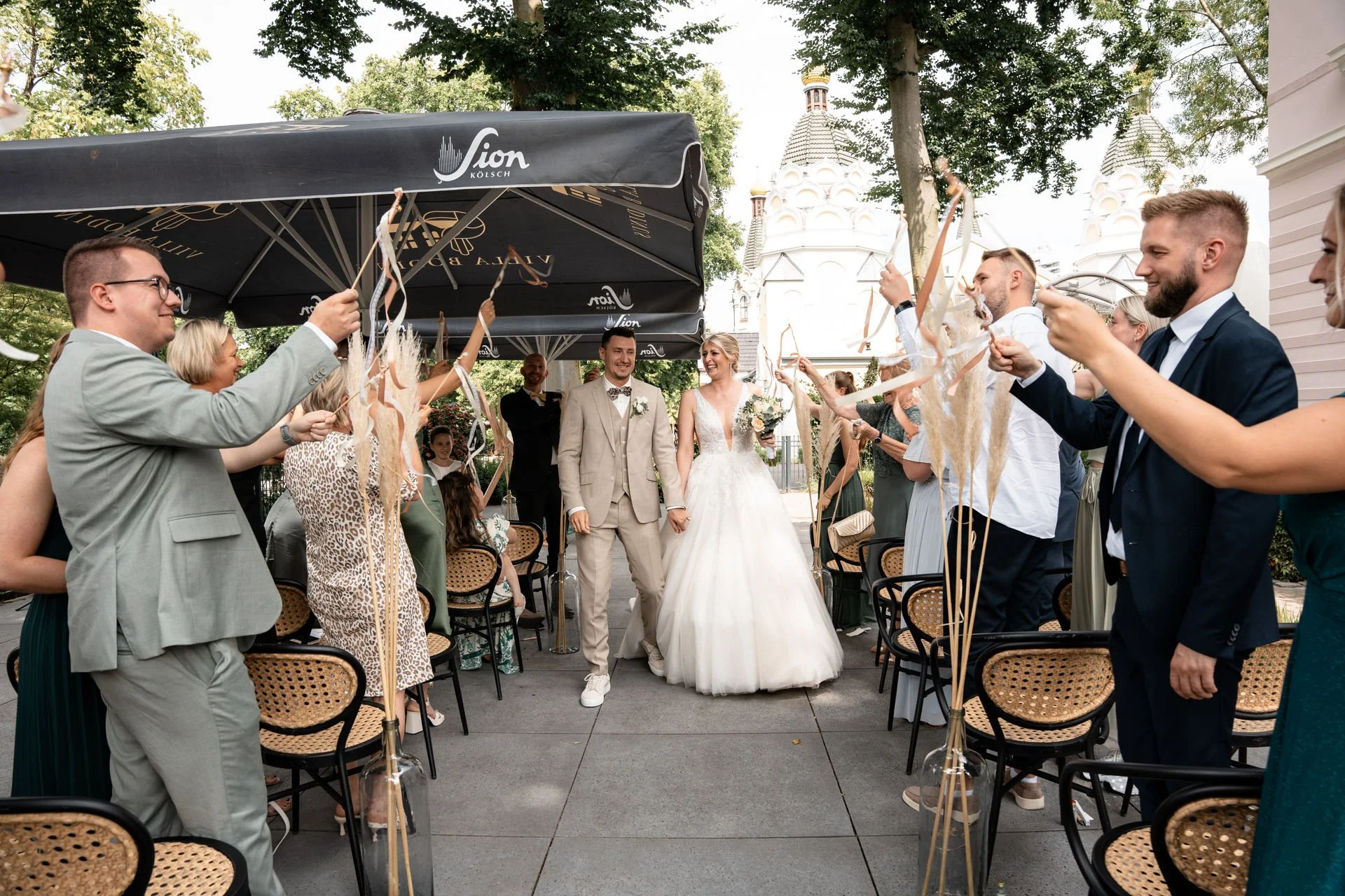 Braut und Bräutigam gehen Hand in Hand durch jubelnde Gäste, die ihre Gästebanner hochhalten, bei einer Hochzeit im Freien, mit weißen Gebäuden im Hintergrund.