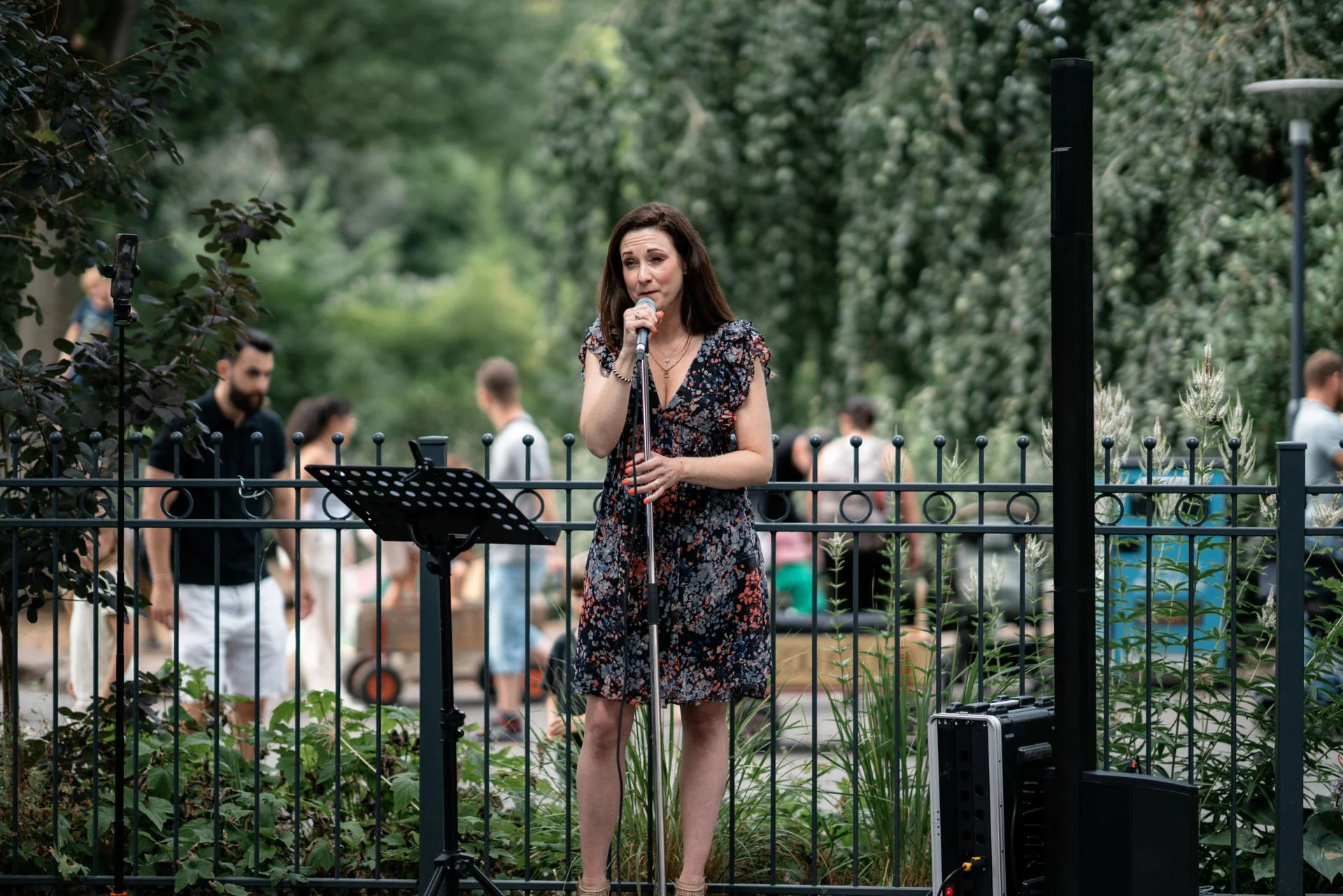 Eine Frau singt bei einem öffentlichen Auftritt in einem Park, umgeben von einem Gitterzaun und Bäumen, mit Publikum im Hintergrund.