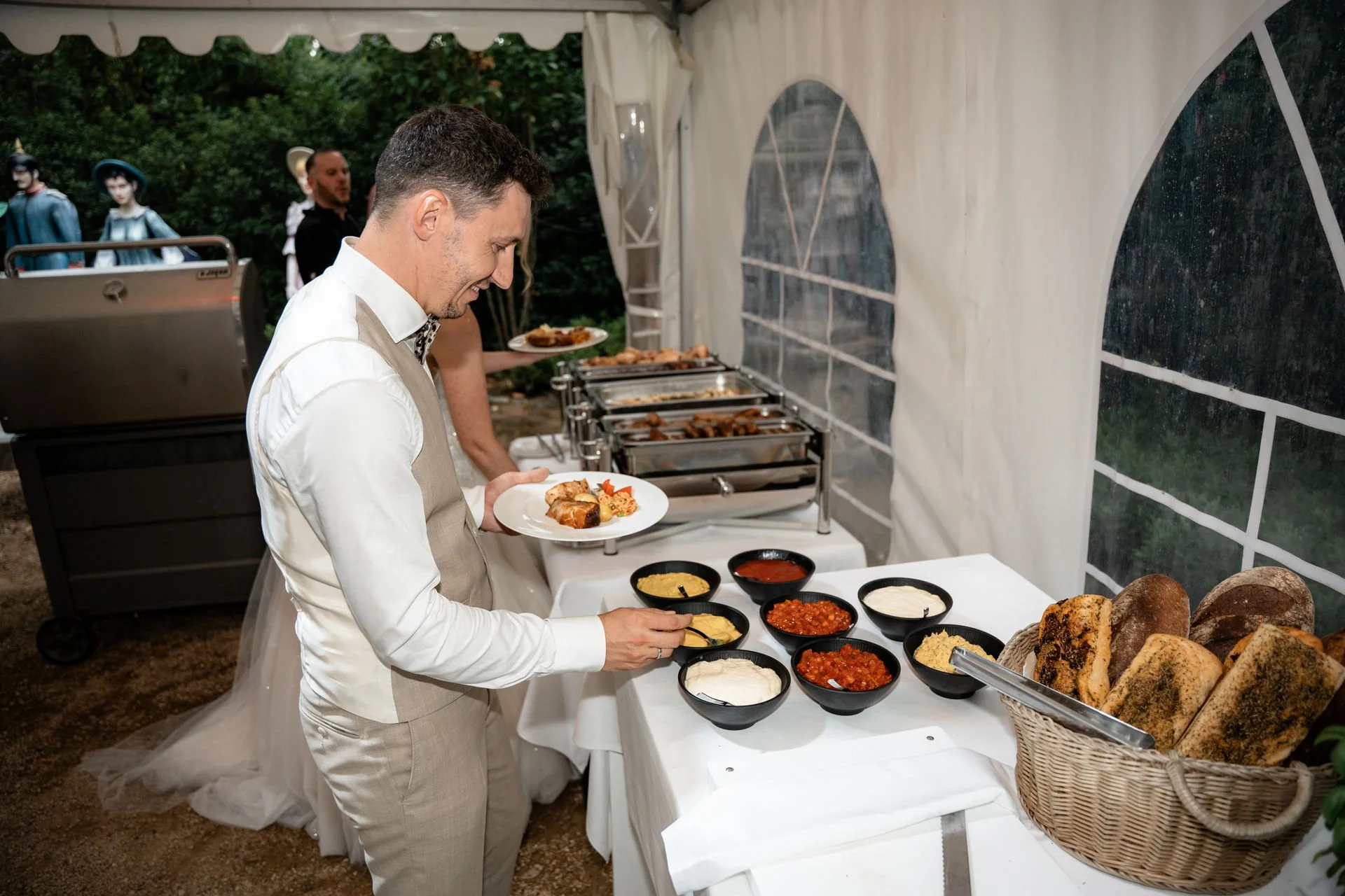 Ein Mann beim Büfett auf einer Party in einem Zelt, serviert sich aufs Teller, im Hintergrund sind weitere Menschen und Dekoration zu sehen.
