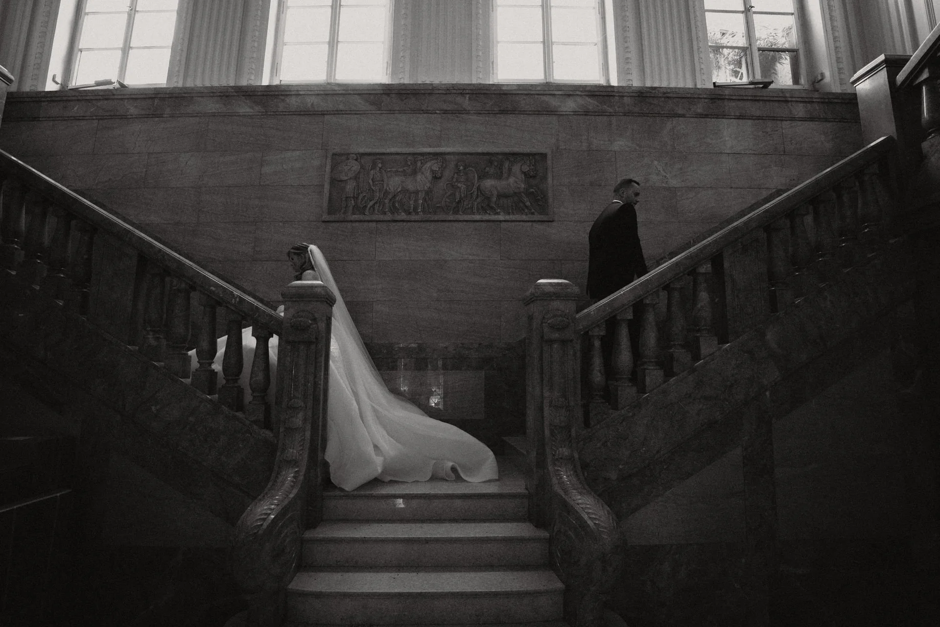 Ein schwarzer und weißer Hochzeitsfotografie eines Brautpaares auf einer hölzernen Treppe in einem alten, eleganten Gebäude mit hohen Fenstern.