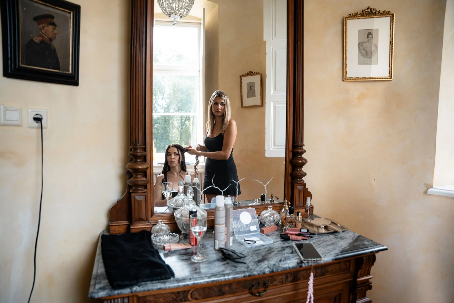 Zwei Frauen, eine sitzend und eine stehend, bei einer Frisörsession im Spiegel eines alten Holzschminktisches. Der Tisch ist voll mit Schönheitsprodukten. Im Hintergrund ein Fenster mit natürlichem Licht.