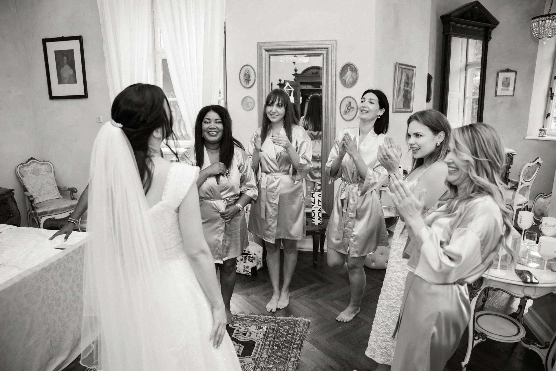 Hochzeitsvorbereitungen: Braut mit fünf Freundinnen im Schlafzimmer, alle lachen und applaudieren in Bademänteln.