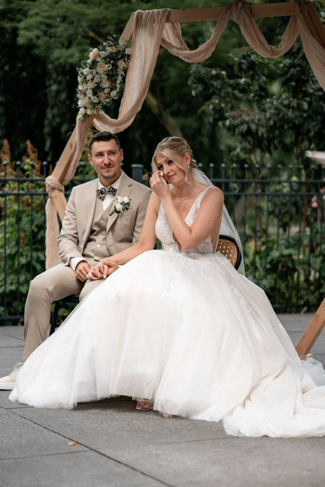 Ein Hochzeitsbild mit einem Brautpaar, das im Freien unter einem dekorativen Holzbogen sitzt. Die Braut trägt ein elegantes weißes Hochzeitskleid und scheint emotional zu sein, während der Bräutigam im beige Anzug lächelt.