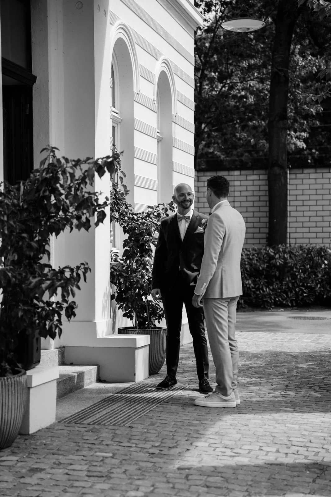 Zwei Männer in formeller Kleidung, die sich draußen vor einem Gebäude unterhalten, mit Bäumen im Hintergrund, in Schwarz-Weiß-Fotografie.