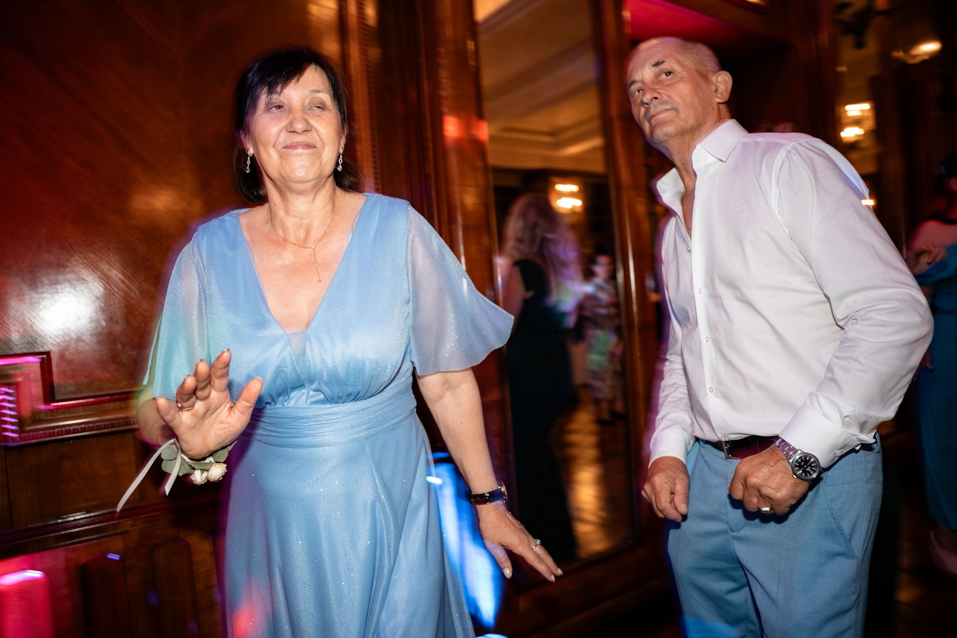 Ein älteres Paar tanzt in einem eleganten Veranstaltungsraum, die Frau trägt ein blaues Kleid und lächelt, während die Hand gehoben wird. Der Mann trägt ein weißes Hemd und blauen Hosen.