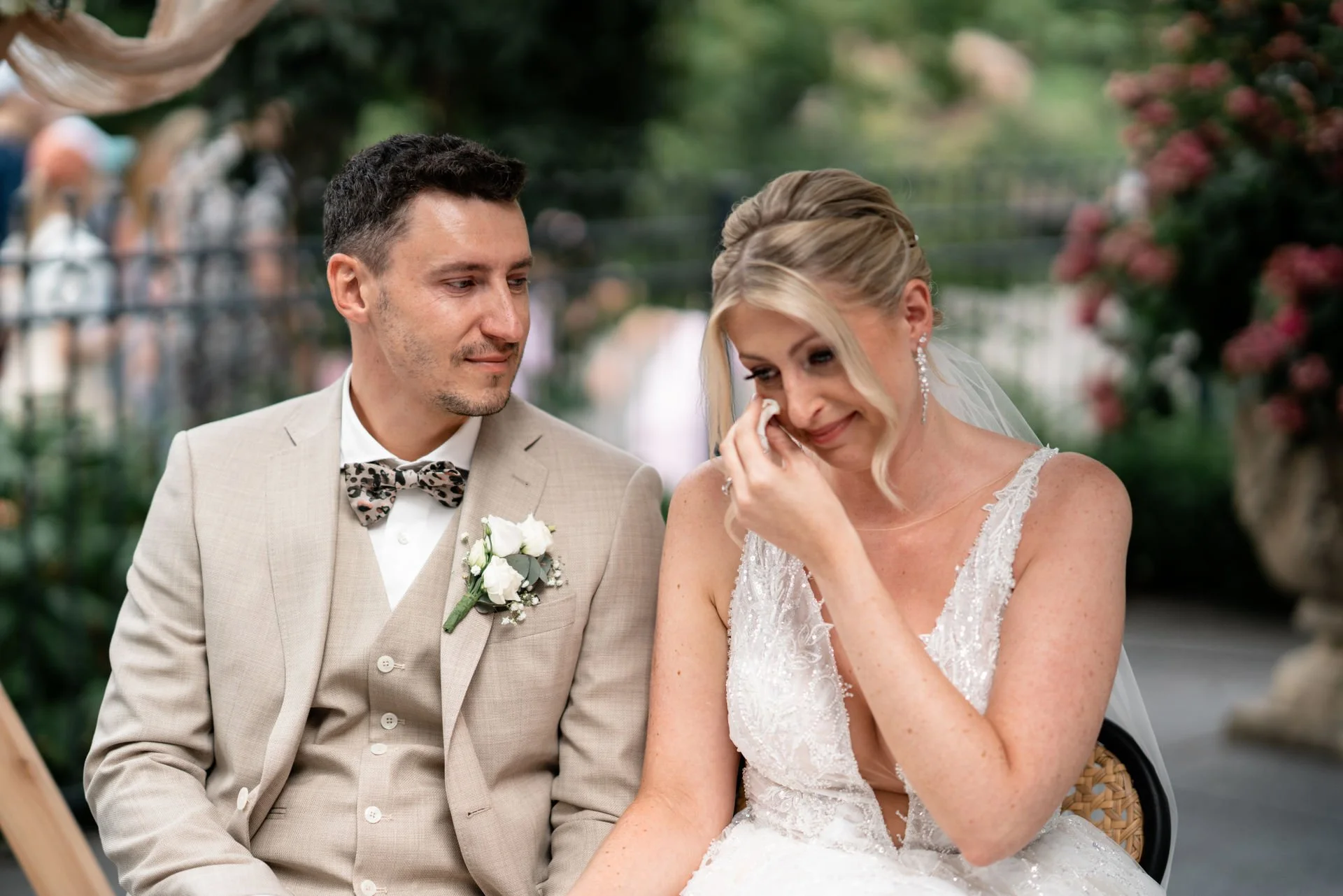 Ein Brautpaar bei einer emotionalen Szene während der Hochzeit im Freien. Die Braut ist am Tränen und hält ein Taschentuch, während der Bräutigam ihr liebevoll zusieht.
