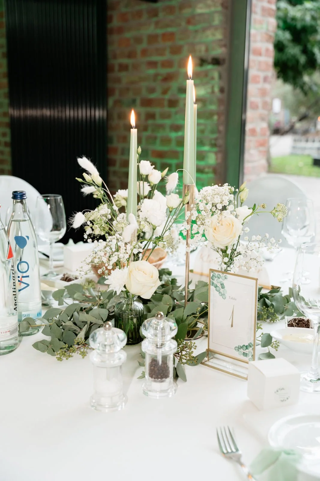 Eleganter gedeckter Tisch mit weißen Blumen, grünen Kerzen, Gläsern und Besteck bei einer Feier oder Hochzeit, im Hintergrund eine Ziegelmauer und Fenster
