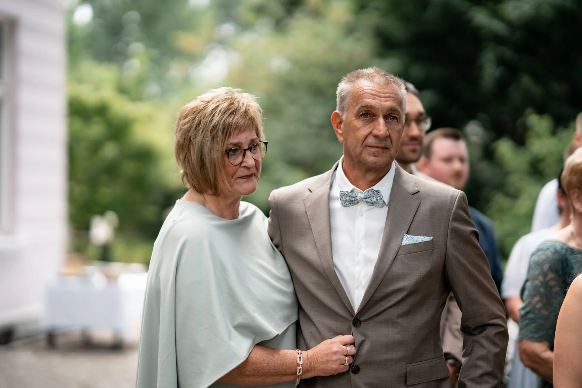 Ein älteres Paar bei einer formellen Veranstaltung im Freien, wahrscheinlich bei einer Hochzeit, mit anderen Gästen im Hintergrund.