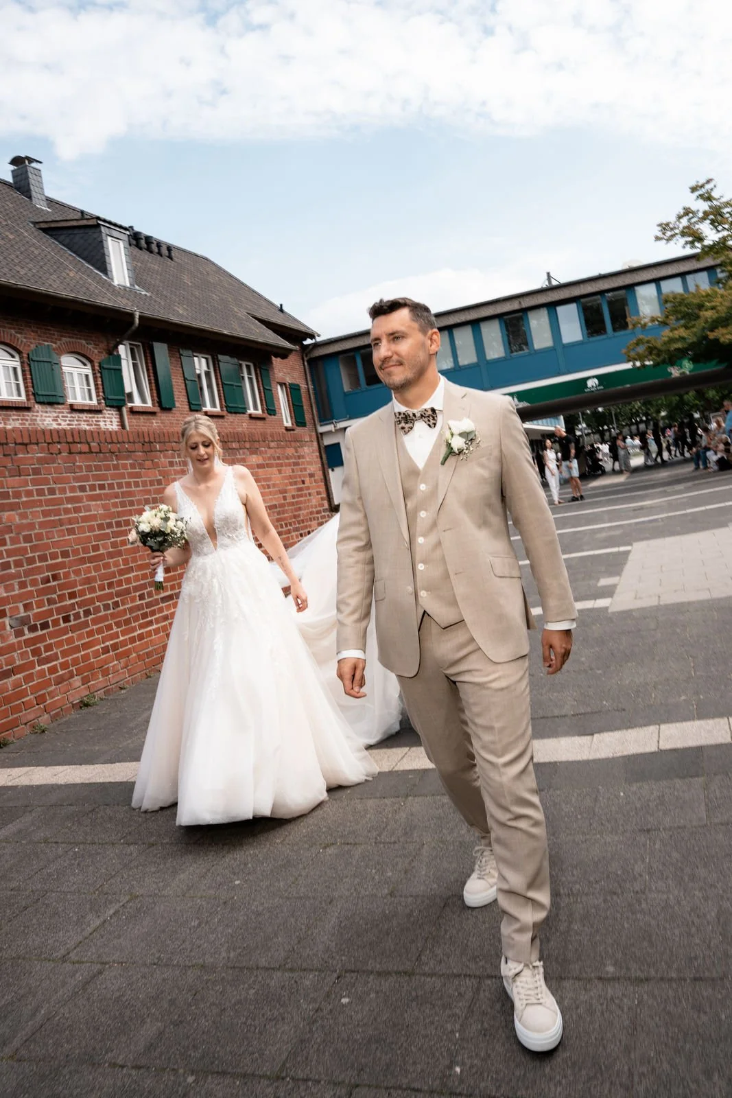 Ein bräunliches Brautpaar geht draußen, der Bräutigam trägt einen hellen Anzug mit Fliege, die Braut trägt ein weißes Kleid und hält einen Blumenstrauß.