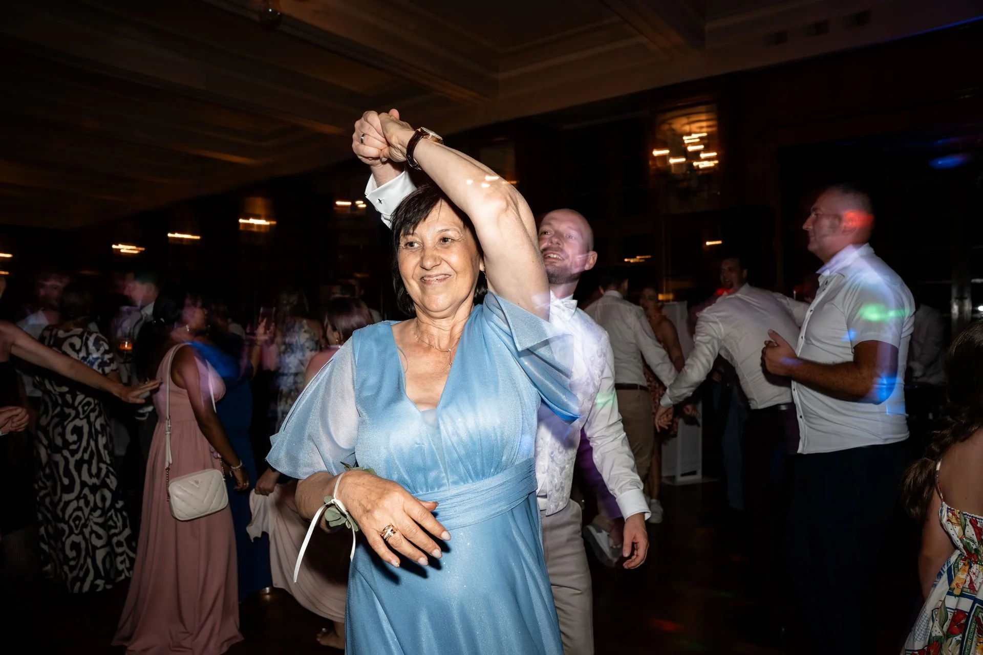 Eine ältere Frau in einem blauen Kleid tanzt mit einem Mann in einem weißen Hemd bei einer Party, im Hintergrund sind weitere tanzende Gäste und bunte Lichter.