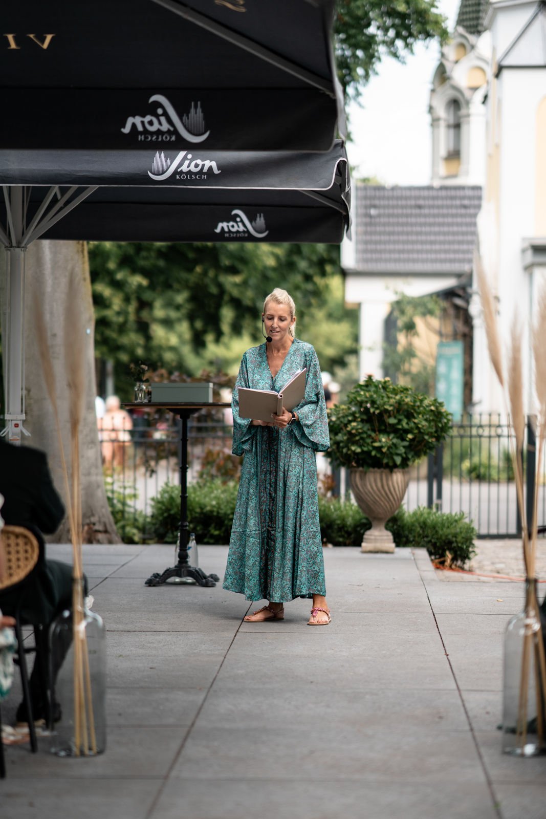 Eine Frau steht auf einer Bühne im Freien und liest aus einem Buch vor. Sie trägt ein langes, gleichmäßiges Kleid mit einem Muster, Sandalen und hat ein Headset. Im Hintergrund sind Bäume und Gebäude sichtbar, es sieht nach einer Veranstaltung im Sta