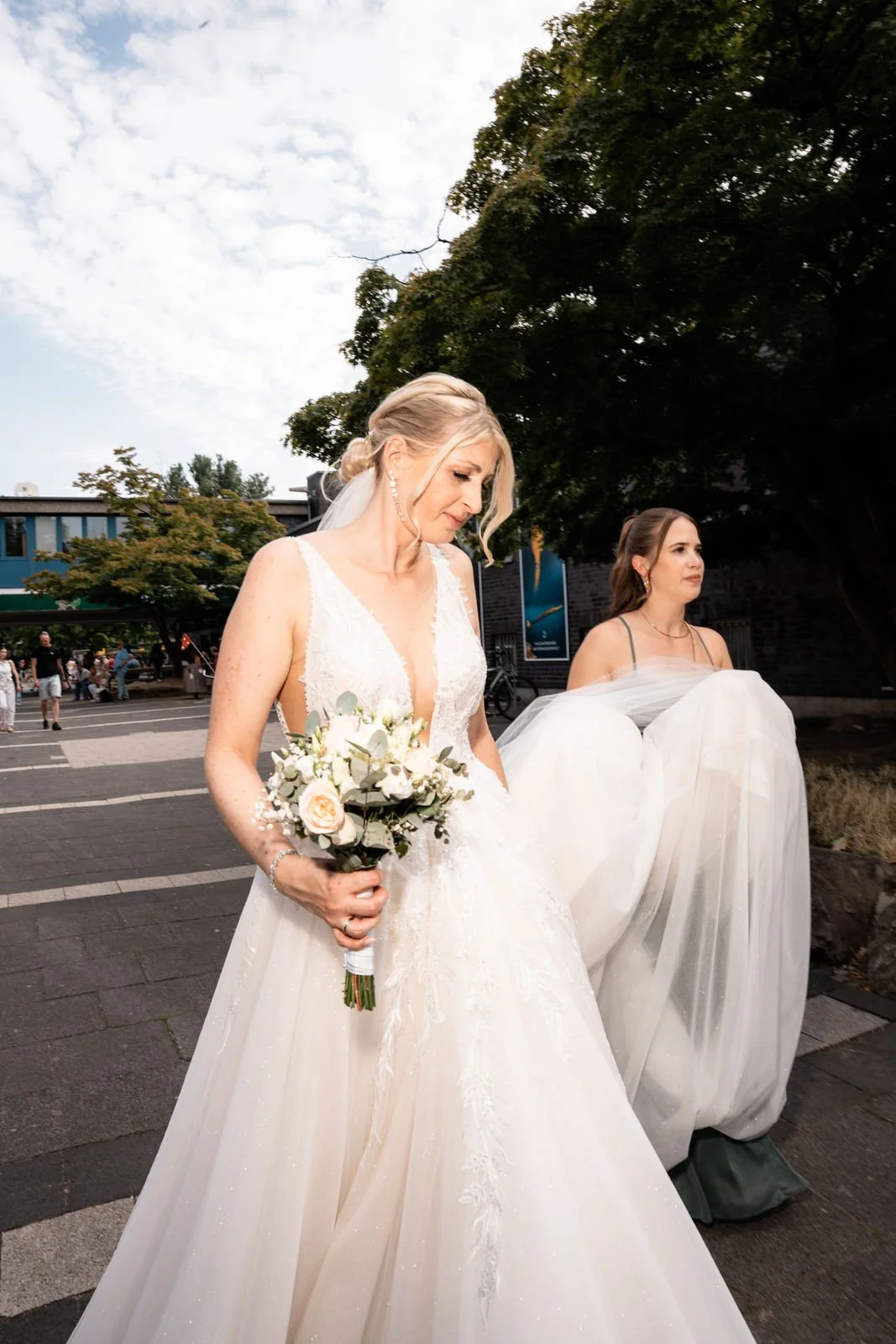 Eine Braut in einem weißen Hochzeitskleid mit einem Blumenstrauß in der Hand, im Freien, während andere Menschen im Hintergrund sind.