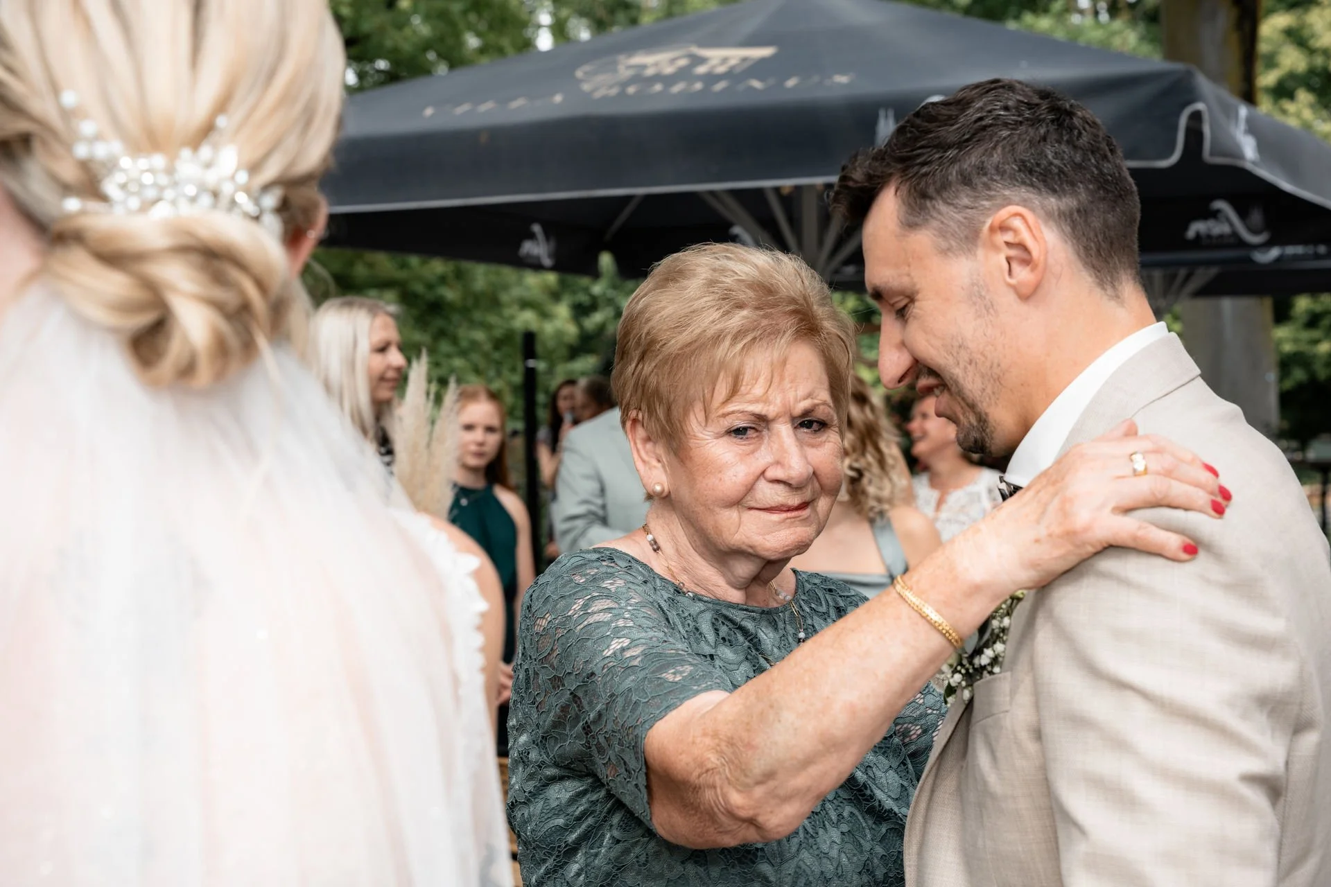 Eine ältere Frau und ein jüngerer Mann bei einer Hochzeitsfeier im Freien, die Frau hält den Mann an den Schultern und beide lächeln. Im Hintergrund sind mehrere Gäste zu sehen.