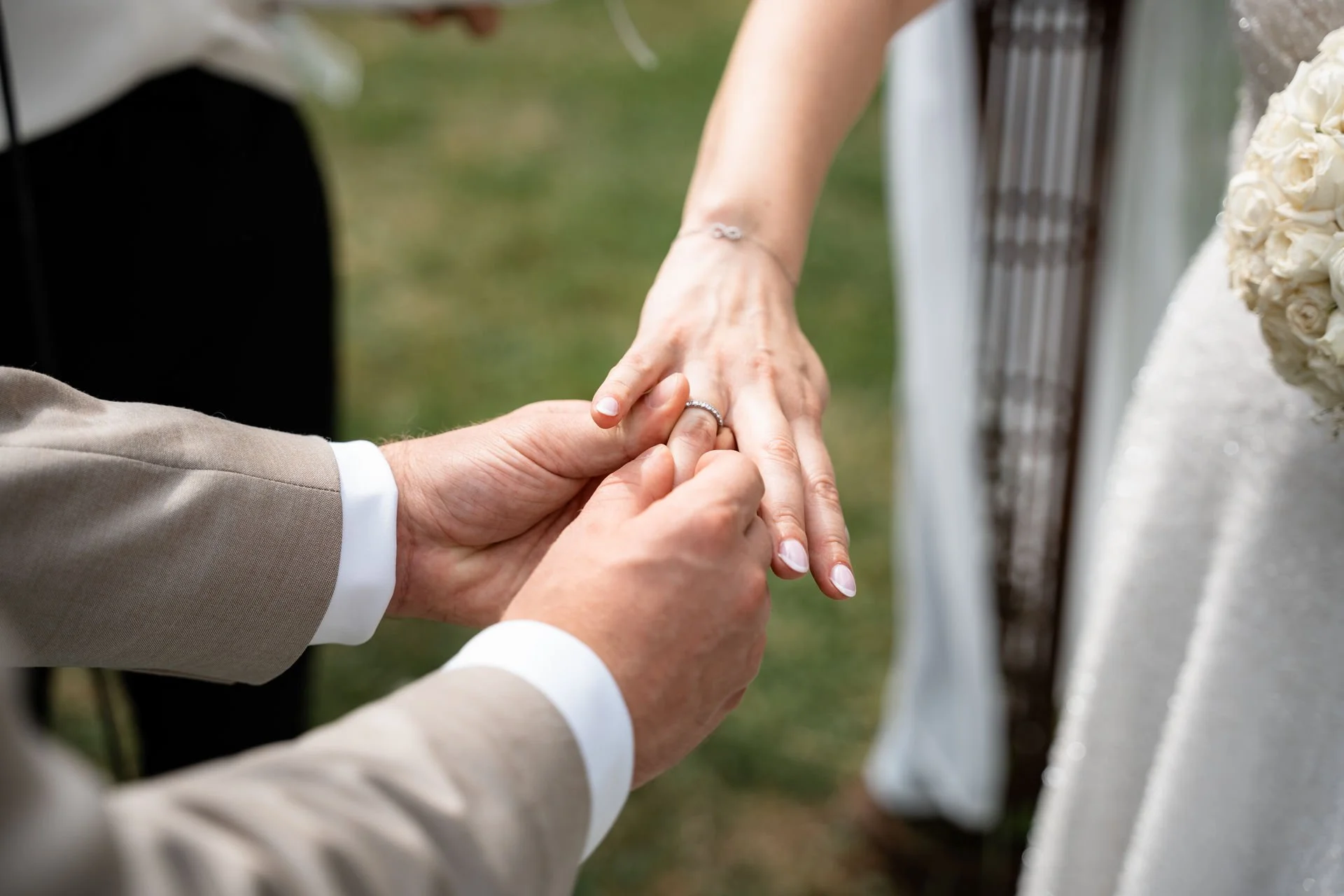 Ein Paar hält sich bei einer Eheschließung an den Händen, die Braut trägt einen Ehering, im Hintergrund steht die Braut in einem weißen Kleid mit Blumenstrauß.