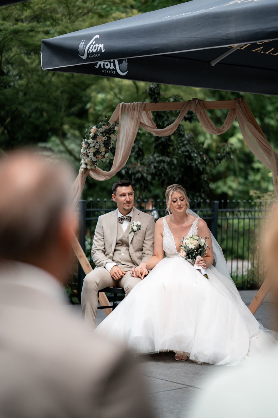 Braut und Bräutigam sitzen bei einer Hochzeit unter einem Holzrahmen mit Tüchern und Blumen, im Hintergrund Bäume und eine Metallzaun, Fotografiert im Freien.