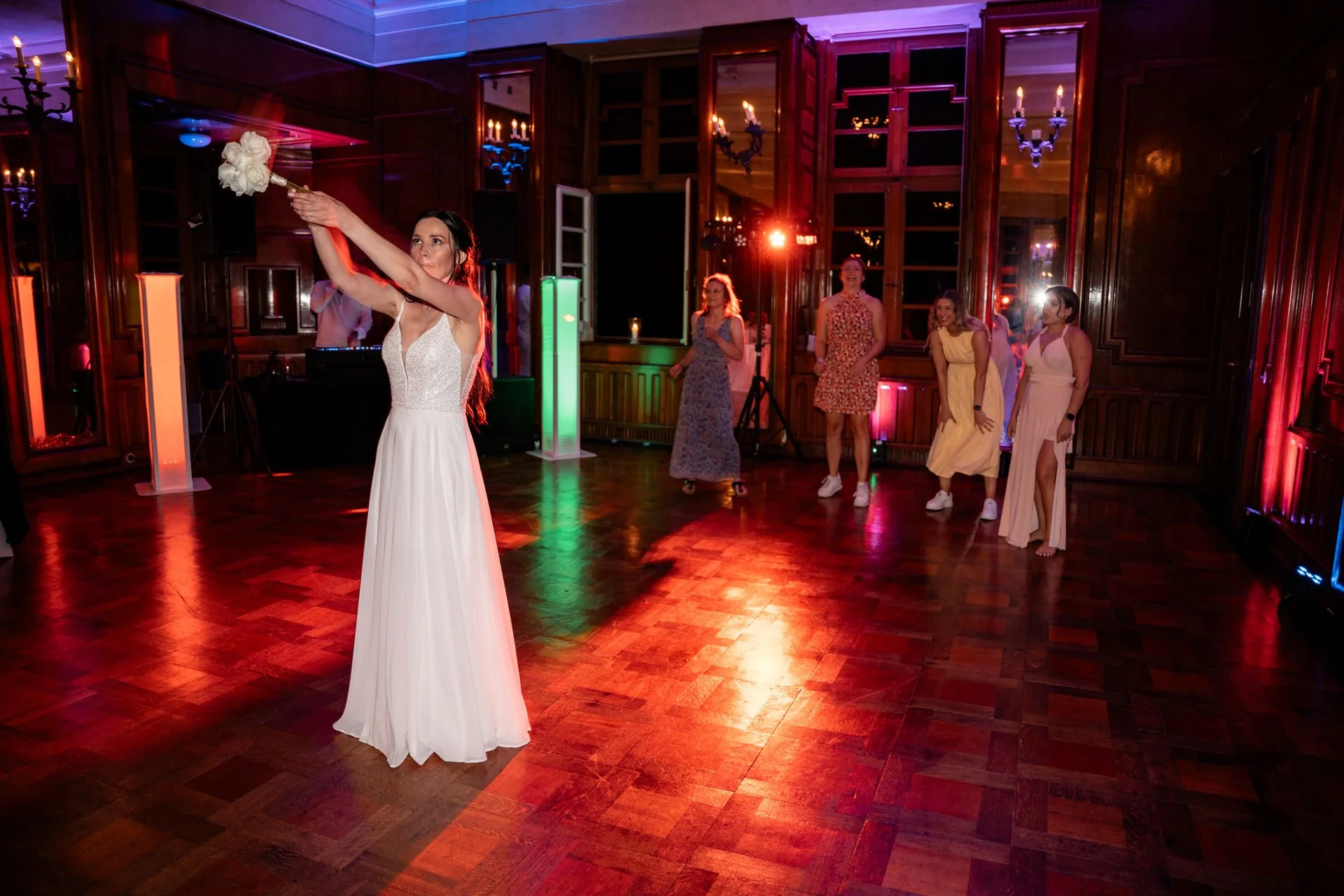 Braut in weißem Kleid wirft Ghose im festlichen Saal mit Holzparkett, umgeben von mehreren lachenden Frauen.