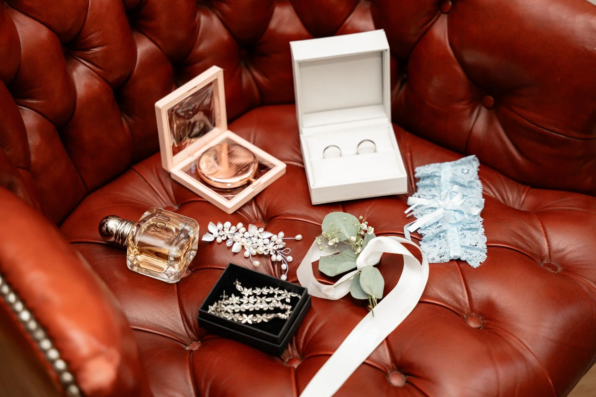 Schmuck und Hochzeitsaccessoires auf einem roten Lederhocker, inklusive Ringe, Parfüm, Schmuckschatullen, Brautschmuck und Blumen.