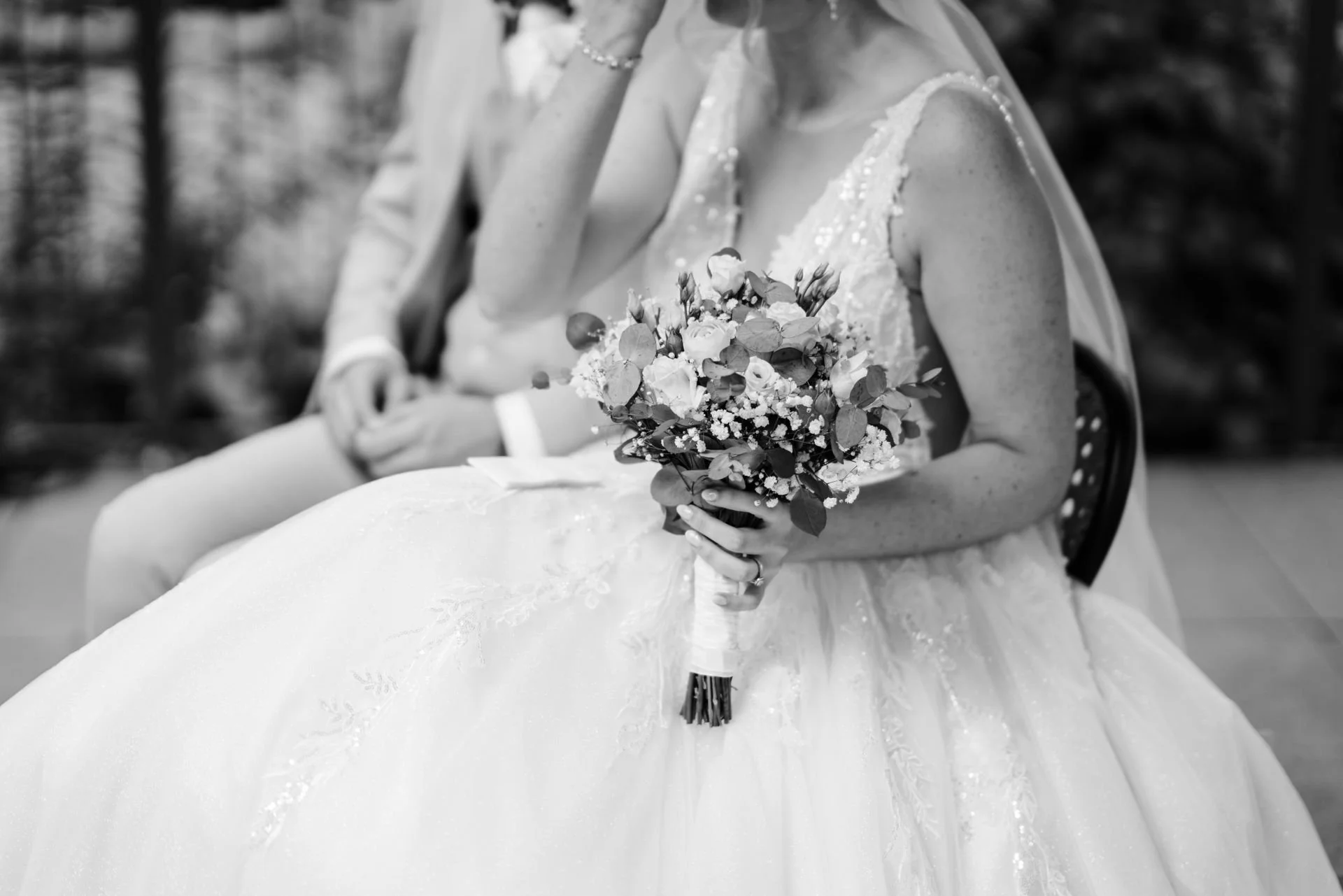 Braut hält einen Blumenstrauß bei einer Hochzeit, im Hintergrund sitzen weitere Personen, alle in formeller Kleidung.