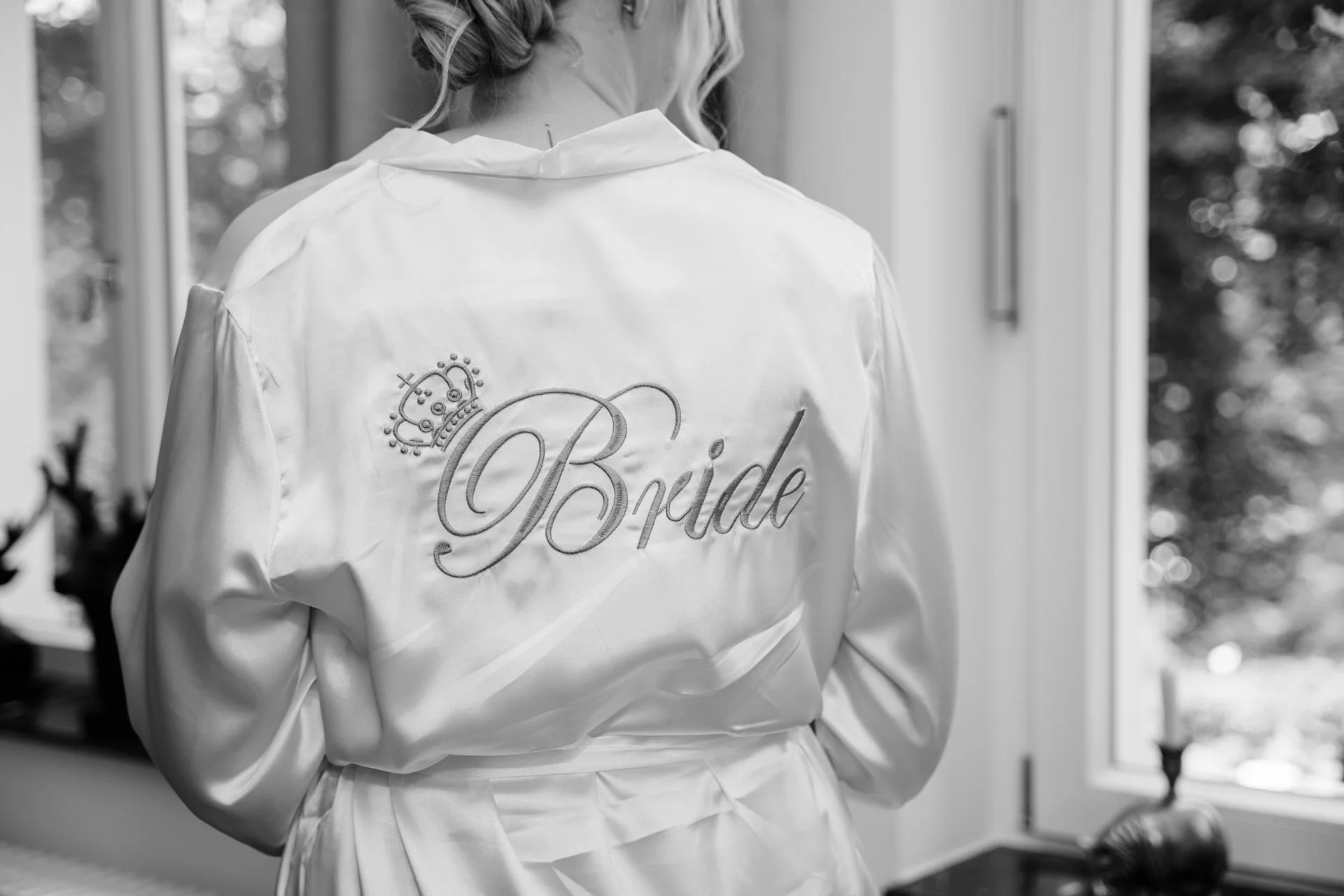 Eine Braut trägt einen weißen Satin-Bademantel mit der Aufschrift 'Bride' und einer Krone. Sie steht vor einem Fenster, im Hintergrund ist ein Kerzenleuchter sichtbar.