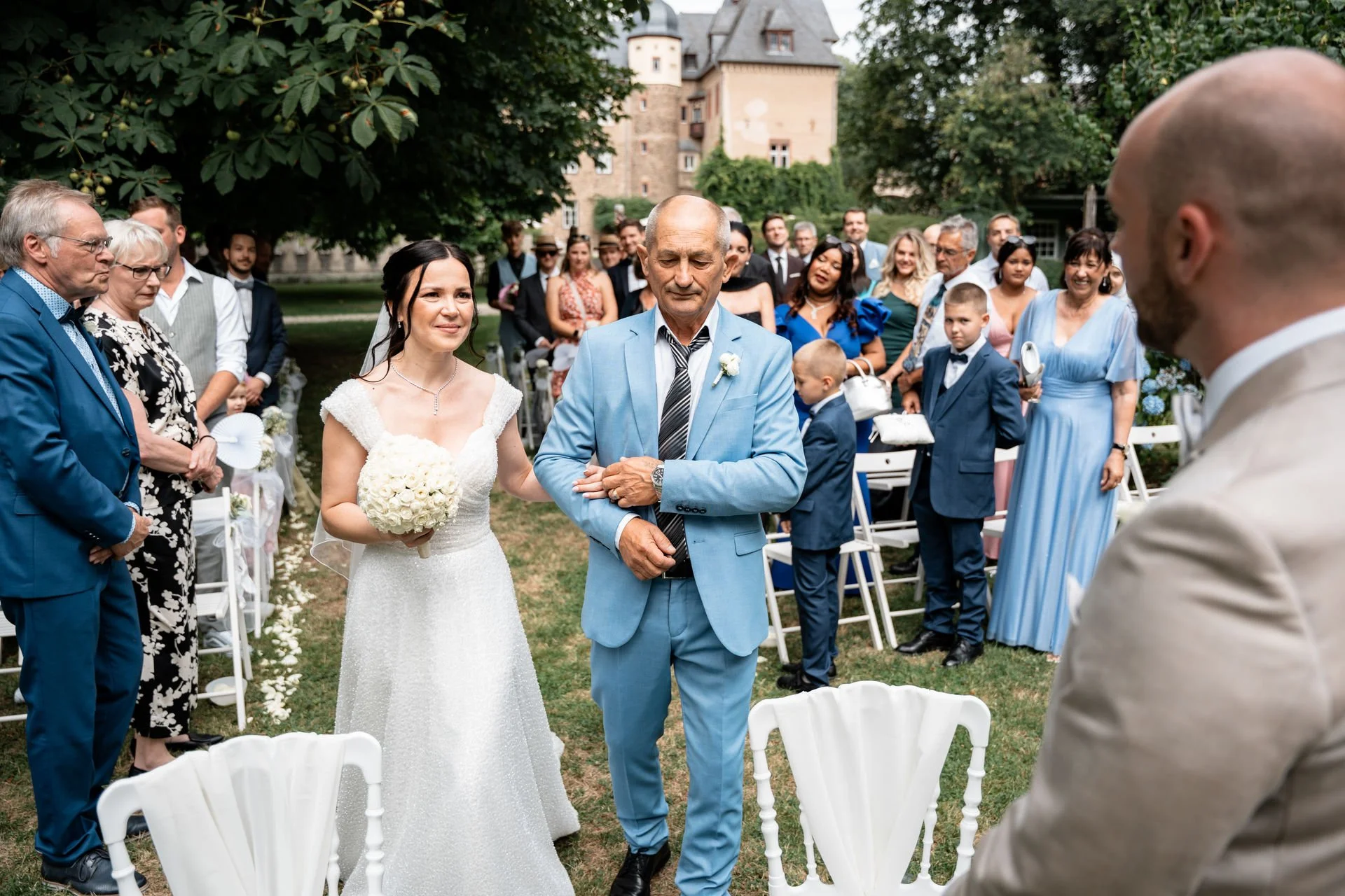 Braut im weißen Kleid hält die Hand eines älteren Mannes in einem hellblauen Anzug während einer Hochzeitszeremonie im Freien, umgeben von Gästen.