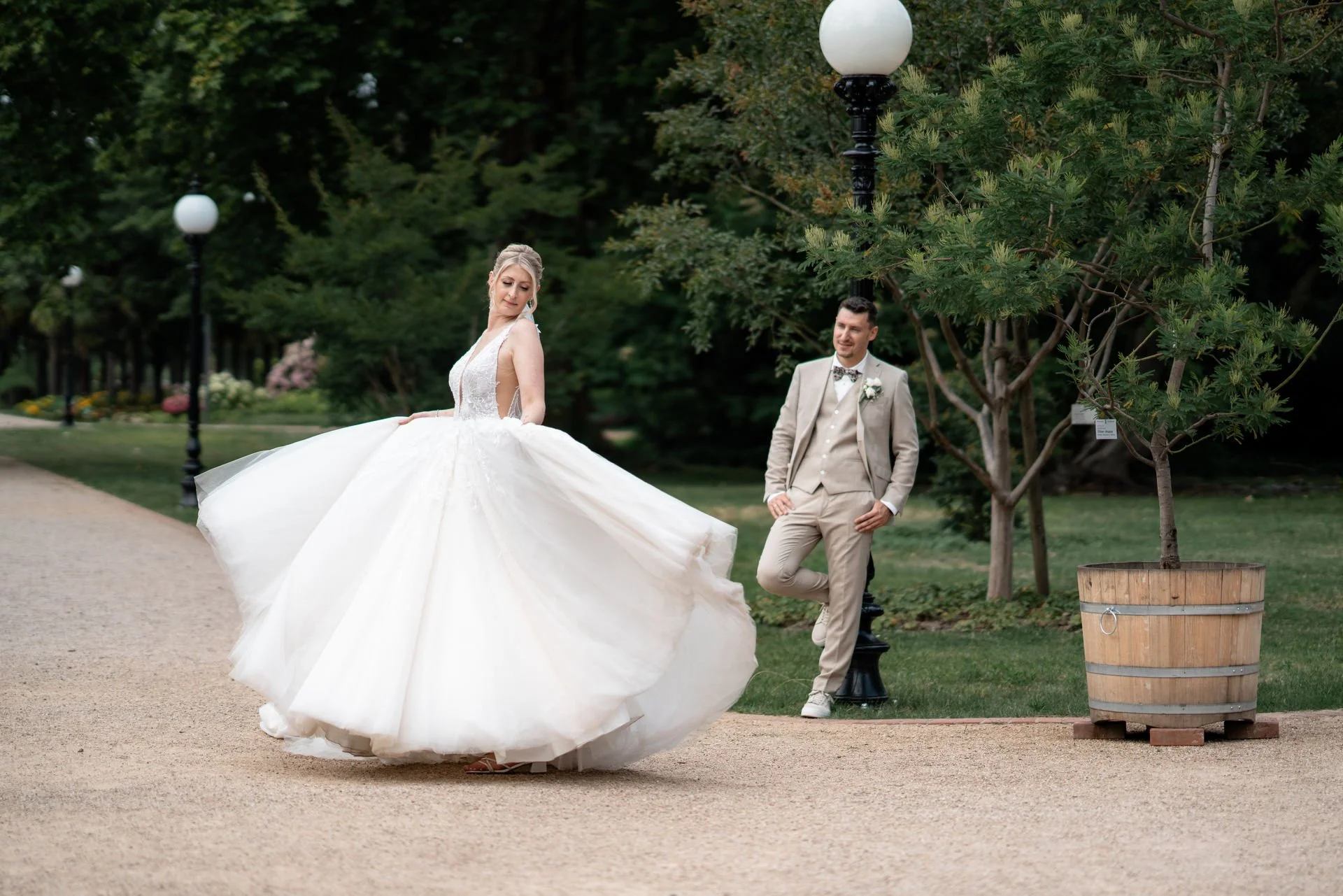 Braut in weißem Hochzeitskleid dreht sich im Park, während der Bräutigam in beige Anzug davor steht und lächelt.