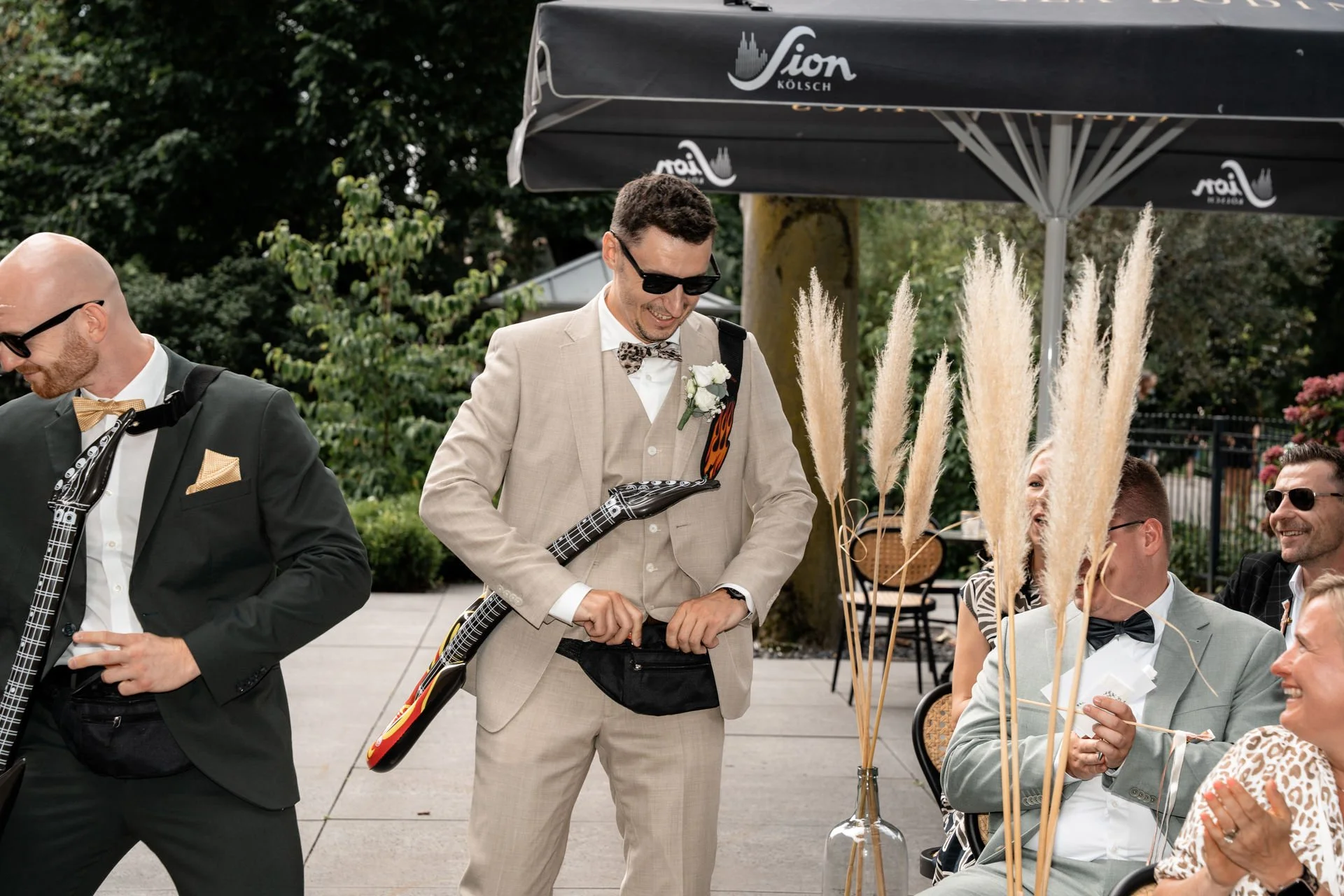 Hochzeitspaar bei der Feier, mit Musikern, die Gitarre spielen, im Freien unter einem Sonnenschirm, mit Blumen, Pflanzen und mehr.