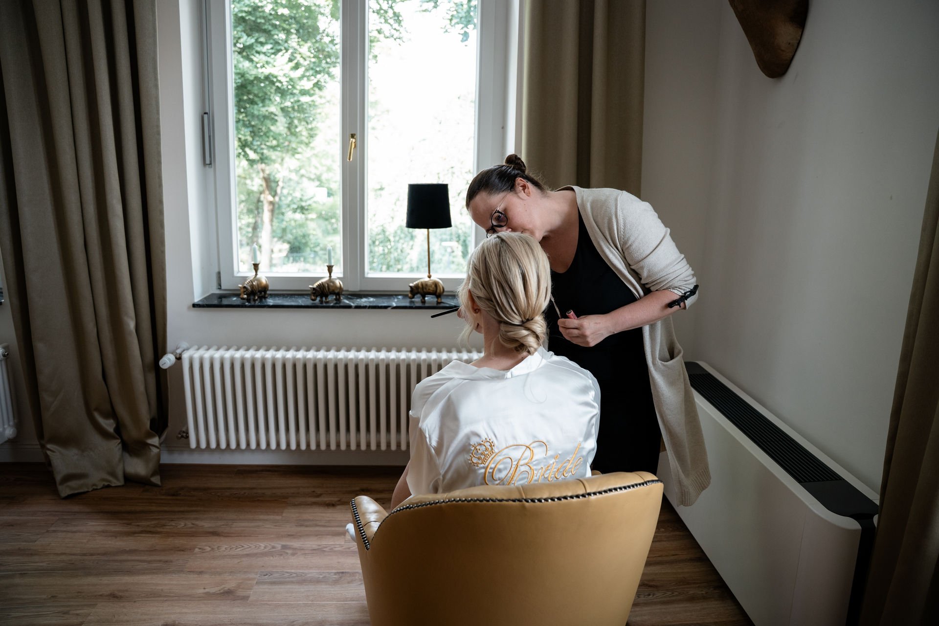 Braut wird von einer Frau geschminkt im Raum mit einem großen Fenster und einem dunklen Lampenschirm auf der Fensterbank