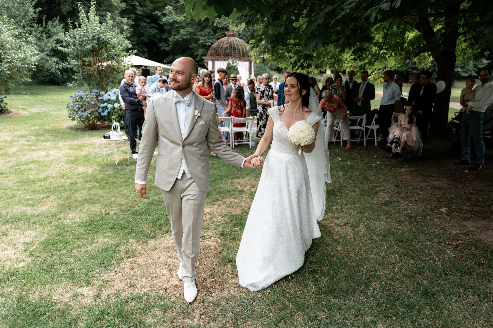 Ein Brautpaar bei ihrer Hochzeit, das Hand in Hand geht, mit Gästen im Hintergrund, unter einem großen Baum auf einer grünen Wiese.