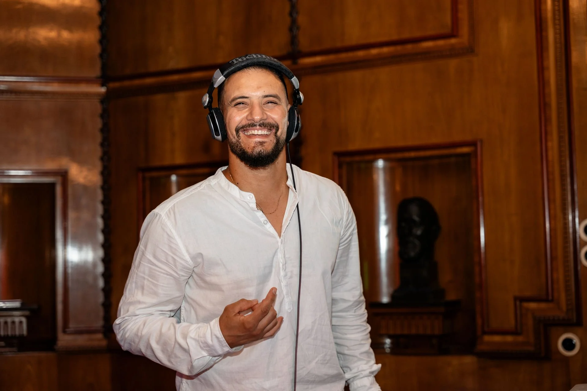 Ein lachender Mann mit dunklem Haar, Bart, in einem weißen Hemd, trägt Kopfhörer, steht in einem Raum mit dunklem, holzvertäfeltem Interieur.
