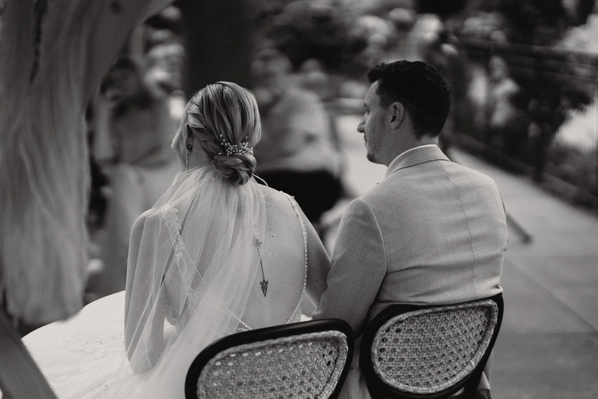 Ein Brautpaar auf ihrer Hochzeitsfeier, sitzend in einem Restaurant, bei einer Zeremonie. Schwarz-weiß-Foto, Perspektive von hinten, Fokus auf die Braut mit Hochsteckfrisur und Schleier und den Bräutigam in Anzug.