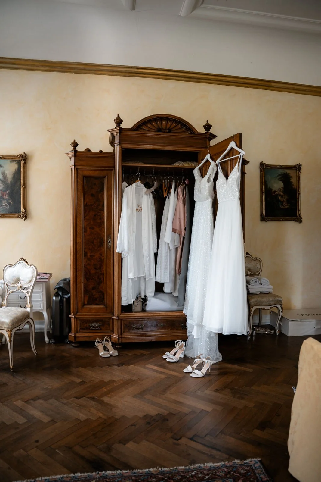 Ein antiker Holzkleiderschrank mit geöffneten Türen, darin hängen Hochzeitskleider und Brautaccessoires. Vor dem Schrank stehen weiße High Heels auf einem dunklen Parkettboden. Der Raum ist elegant und traditionell eingerichtet, mit Wandkunst und Vin