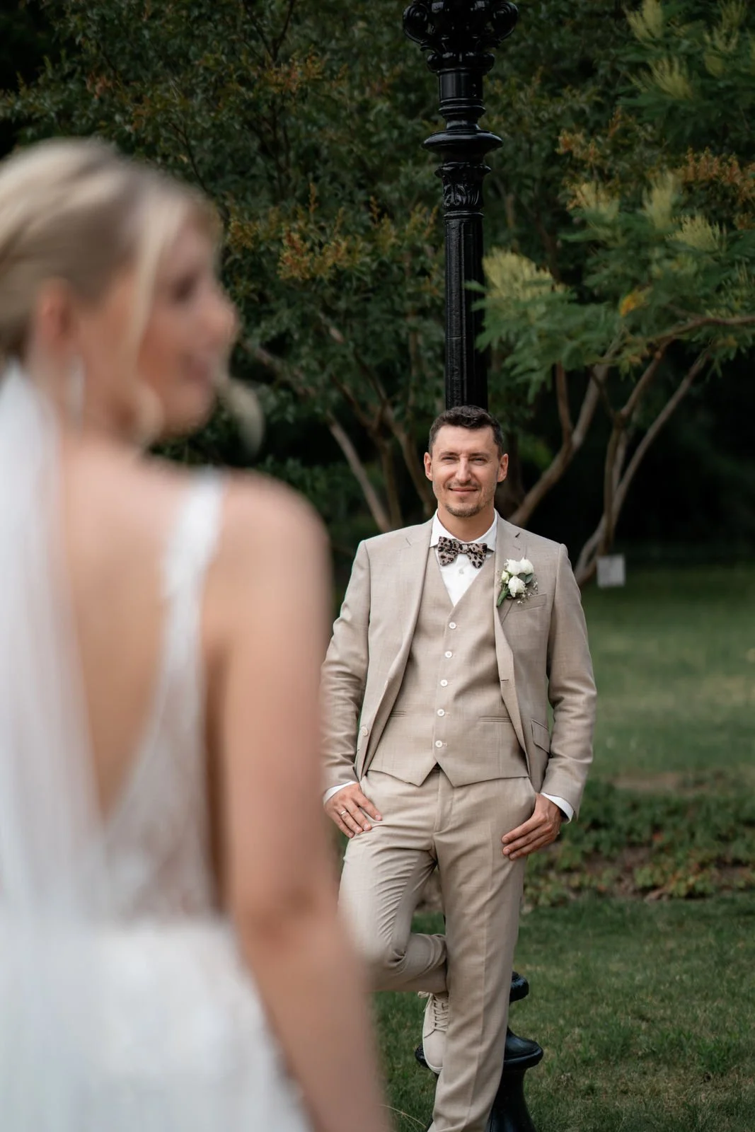 Ein bräutiger Mann in einem beige Anzug mit Weste und Fliege, der an einem Park oder Garten steht, während eine Braut im Vordergrund unscharf betrachtet wird.