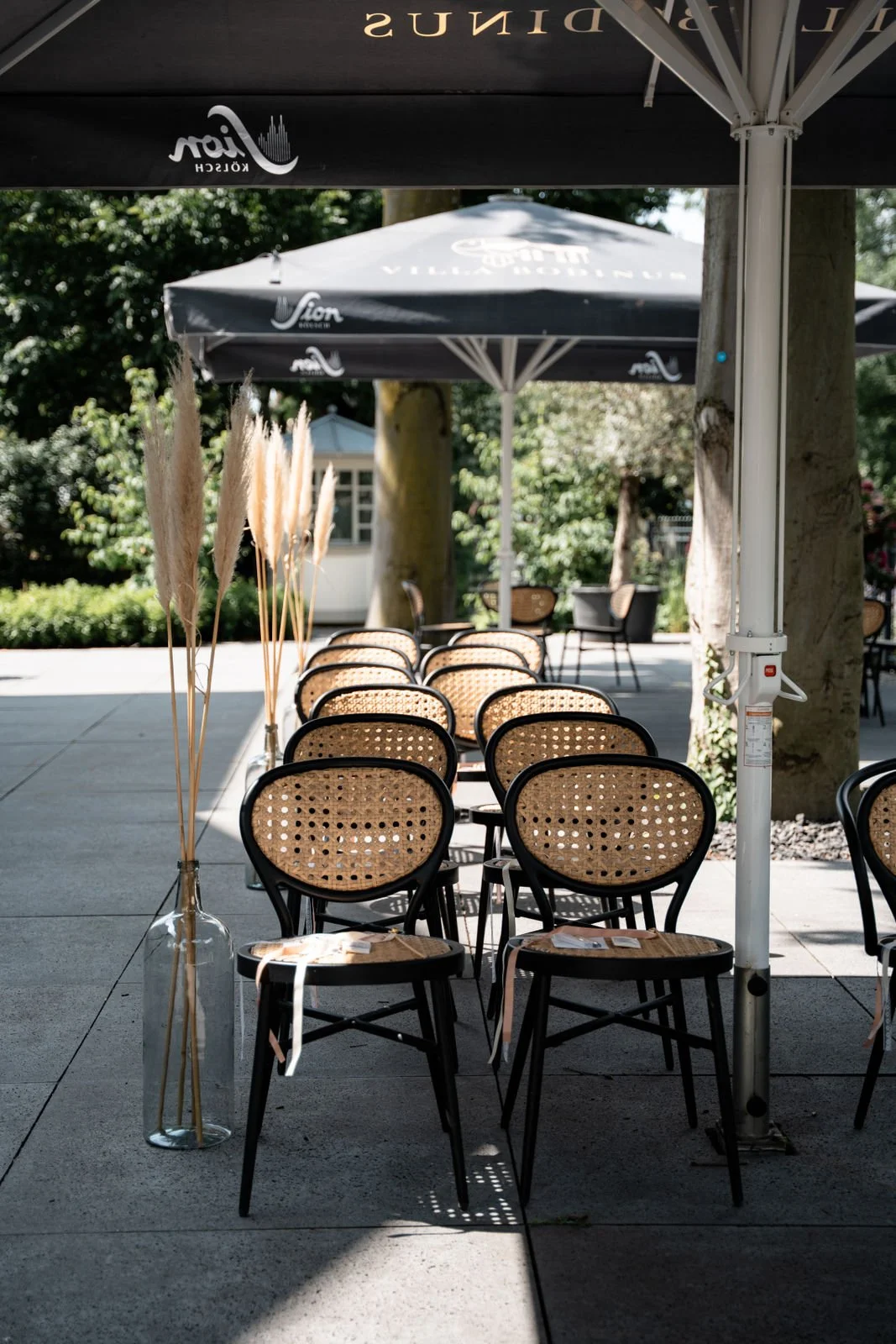 Leere schattige Terrasse im Freien mit gestapelten Stühlen, einem großen Sonnenschirm, Dekorationen mit getrockneten Pflanzen in einer Vase, umgeben von Bäumen und Grünflächen.