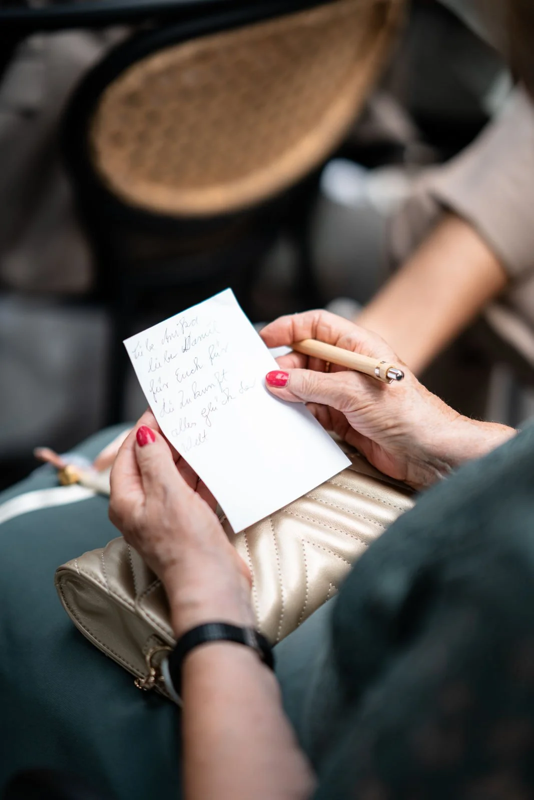 Eine Frau mit roten Nägeln hält einen Stift und schreibt eine handgeschriebene Notiz auf einem weißen Zettel. Sie sitzt in einem öffentlichen Raum, möglicherweise im Zug oder Flugzeug, vor einem Rucksack oder Tasche.