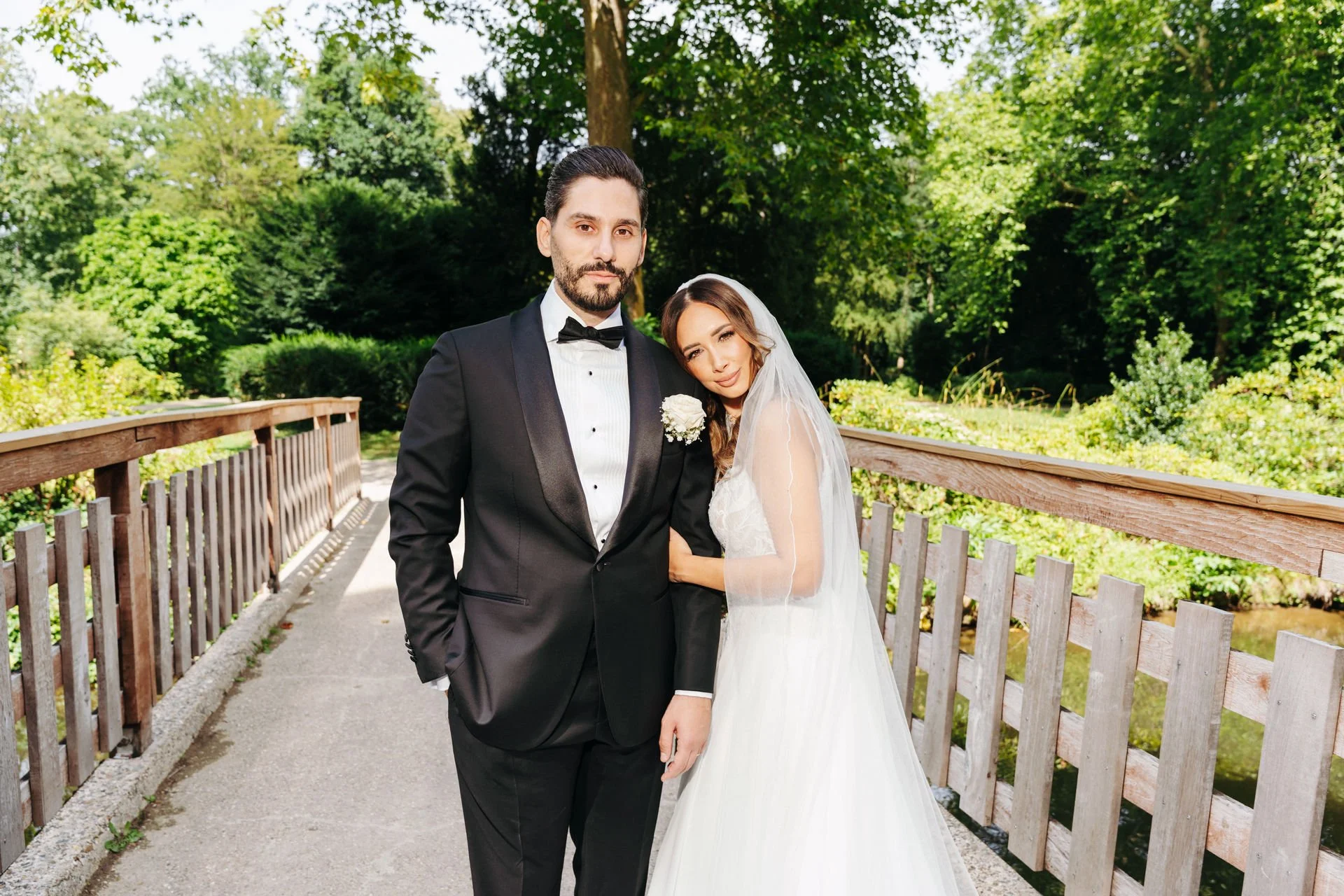 Ein Brautpaar steht im Freien auf einer Brücke, umgeben von grünen Bäumen, das Mädchen trägt ein weißes Brautkleid und einen Schleier, der Mann trägt einen schwarzen Anzug mit Fliege.