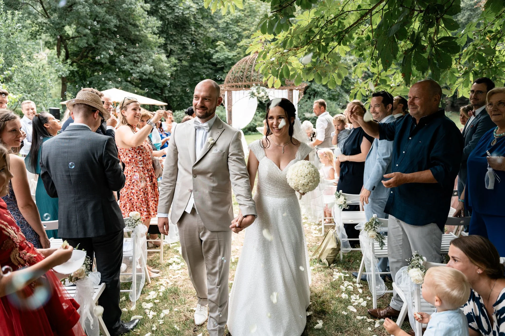 Ein Brautpaar geht bei einer Hochzeit unter einem Baum. Die Braut trägt ein weißes Kleid und hält einen Blumenstrauß, der Bräutigam trägt einen hellen Anzug. Gäste umgeben sie und feiern, während Seifenblasen in der Luft fliegen.